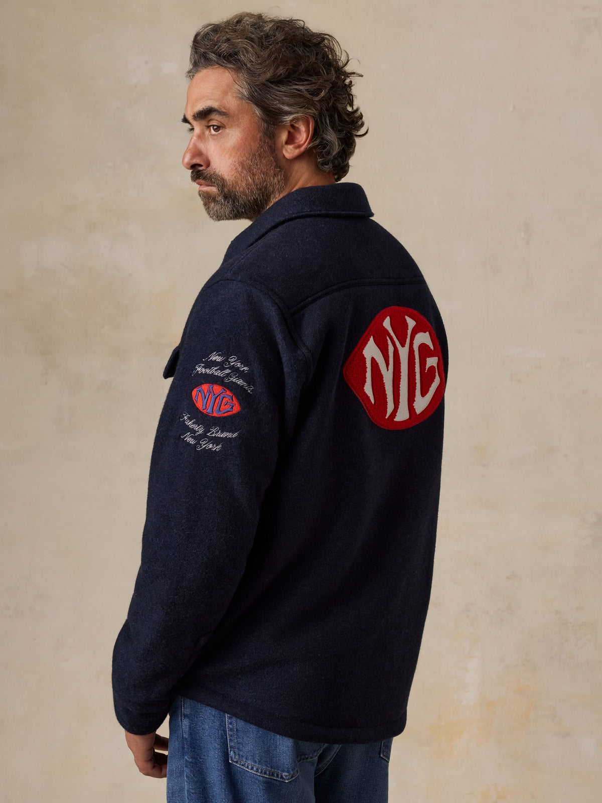 A man viewed from behind wearing a blue NY Giants jacket and blue jeans, image 5 of 5