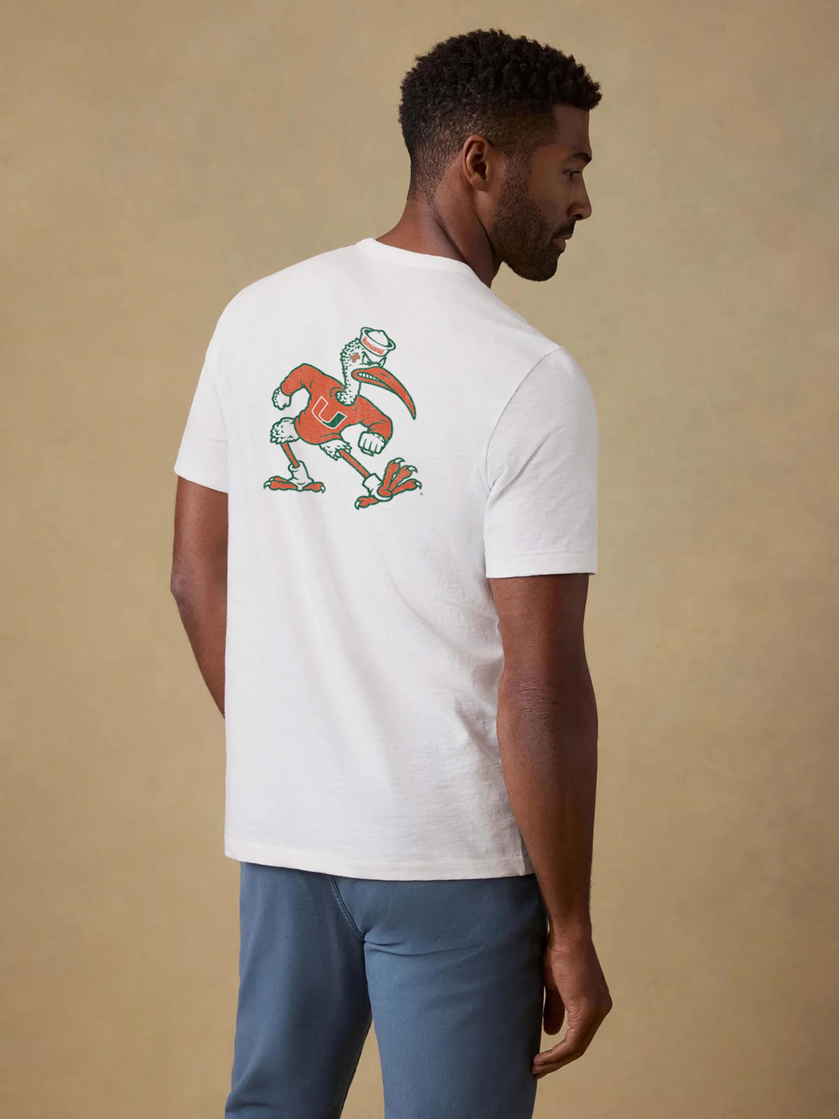 A man viewed from behind wearing a white University of Miami tee and blue pants, image 2 of 2