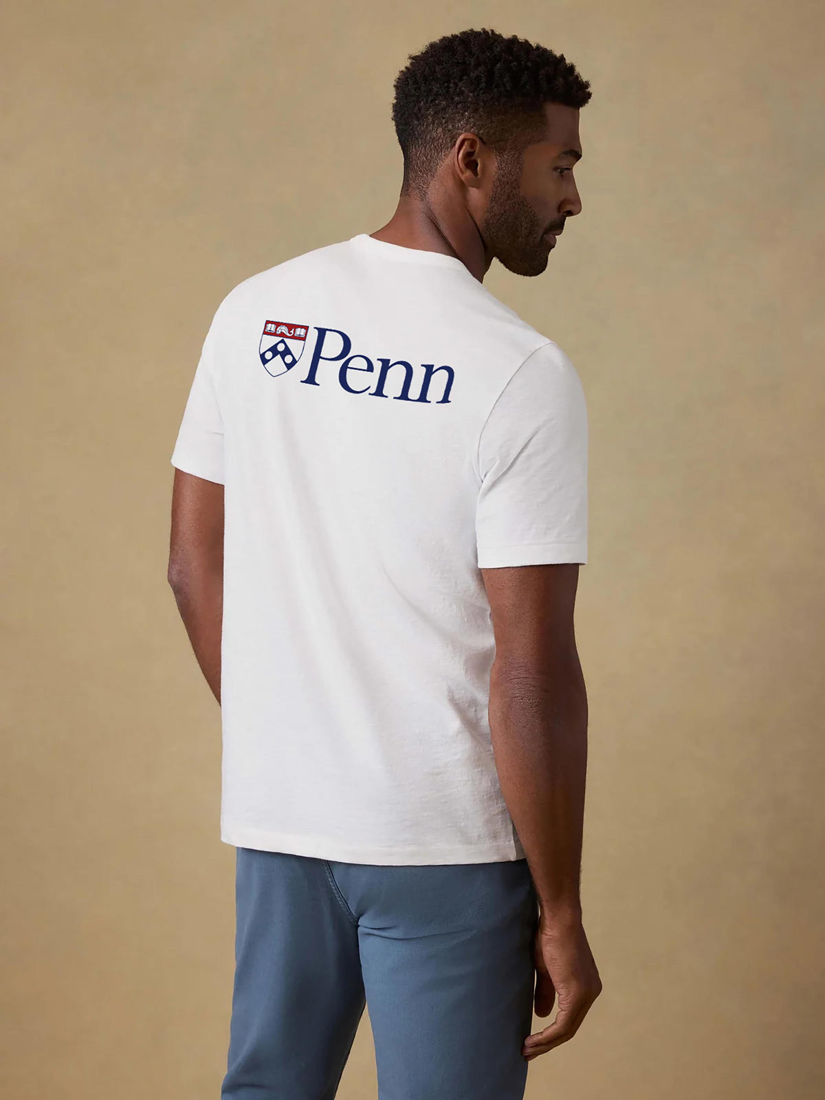 A man viewed from behind wearing a white University of Pennsylvania tee and blue pants, image 2 of 2