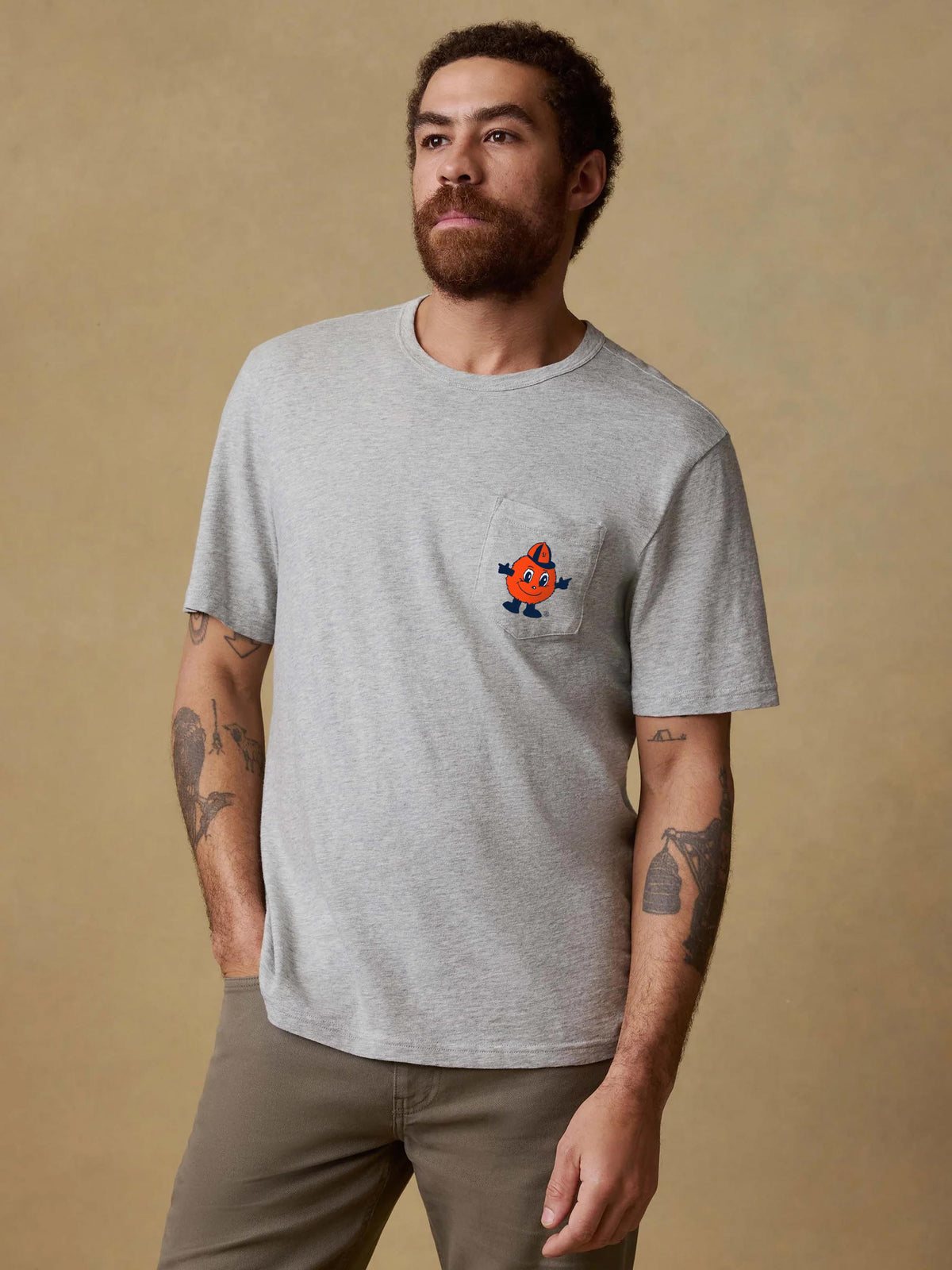 A man wearing a grey Syracuse pocket tee and brown pants, image 1 of 2