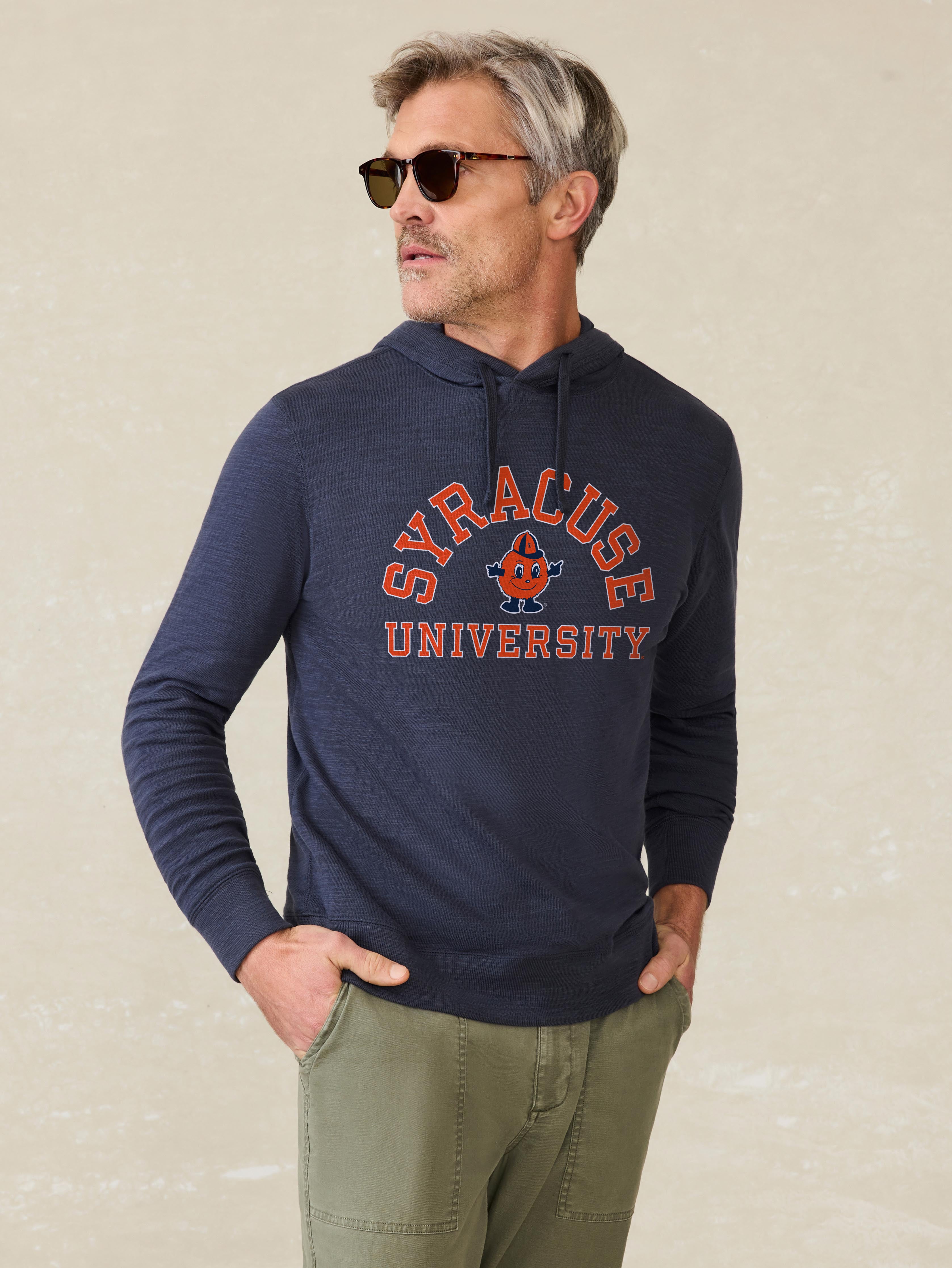 Man is wearing a navy hoodie with Syracuse University graphic on the center chest against a beige background