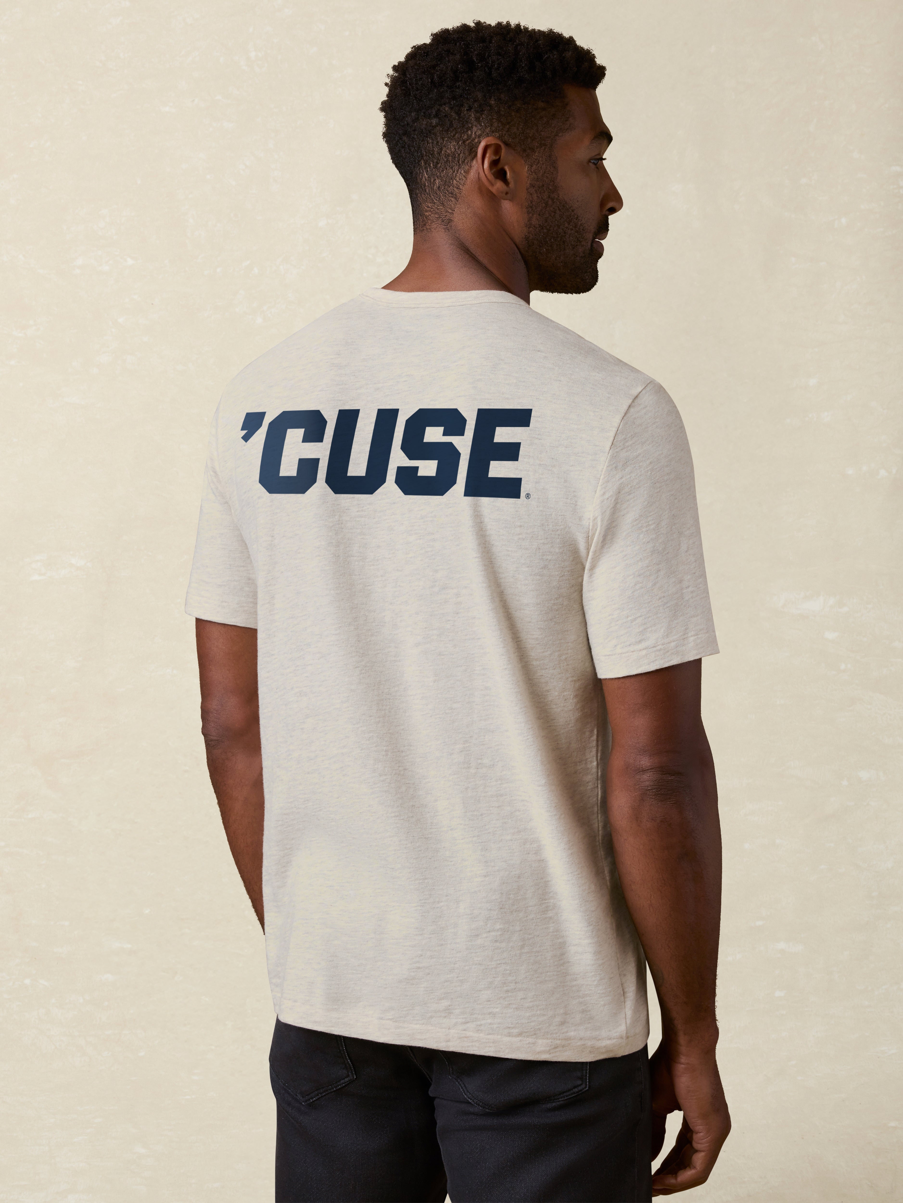 Man wearing a heather grey t-shirt with 'Cuse' printed on the back against a beige background