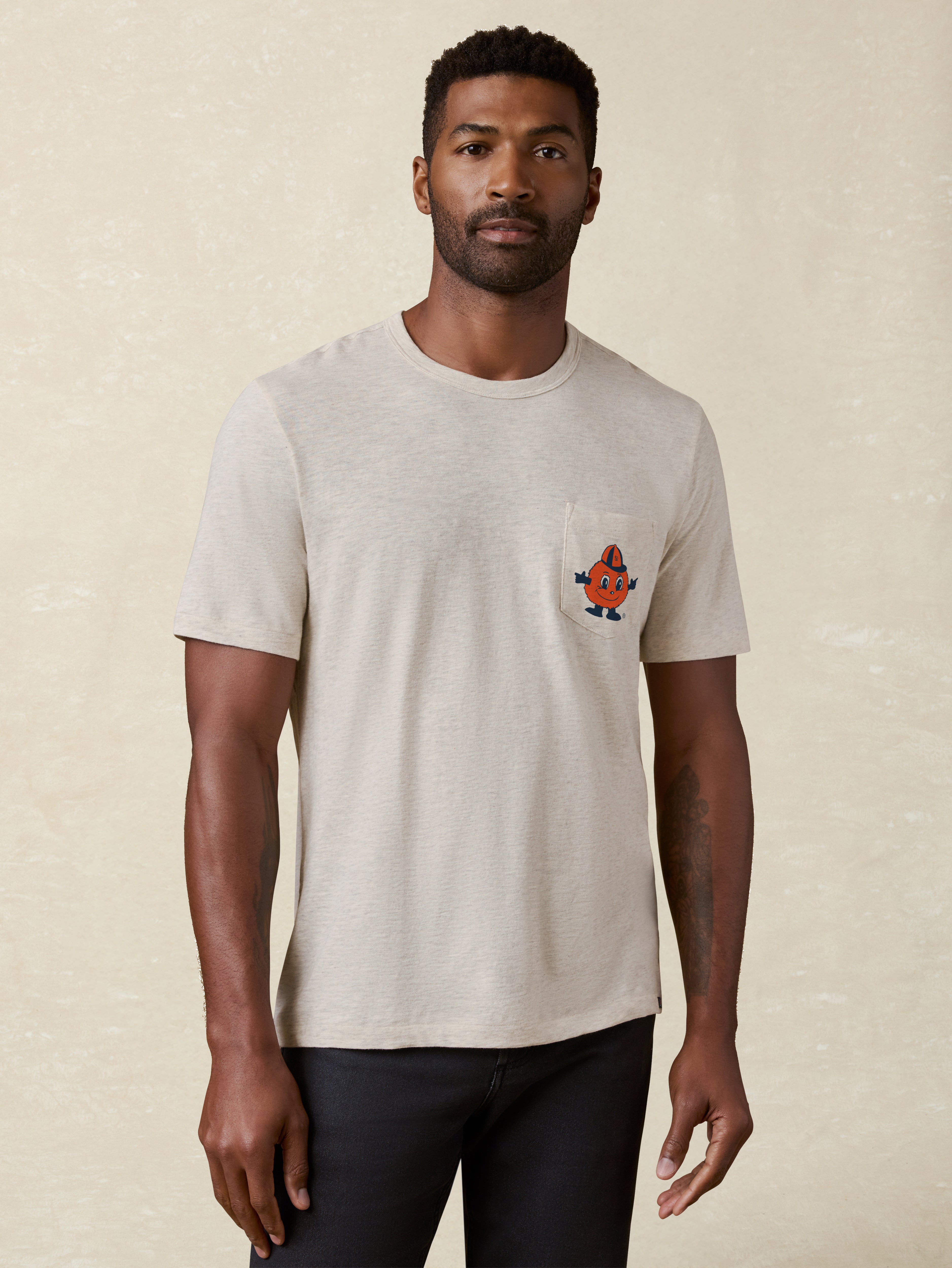 Man wearing a heather grey t-shirt with a Syracuse University graphic on the left chest pocket against a beige background