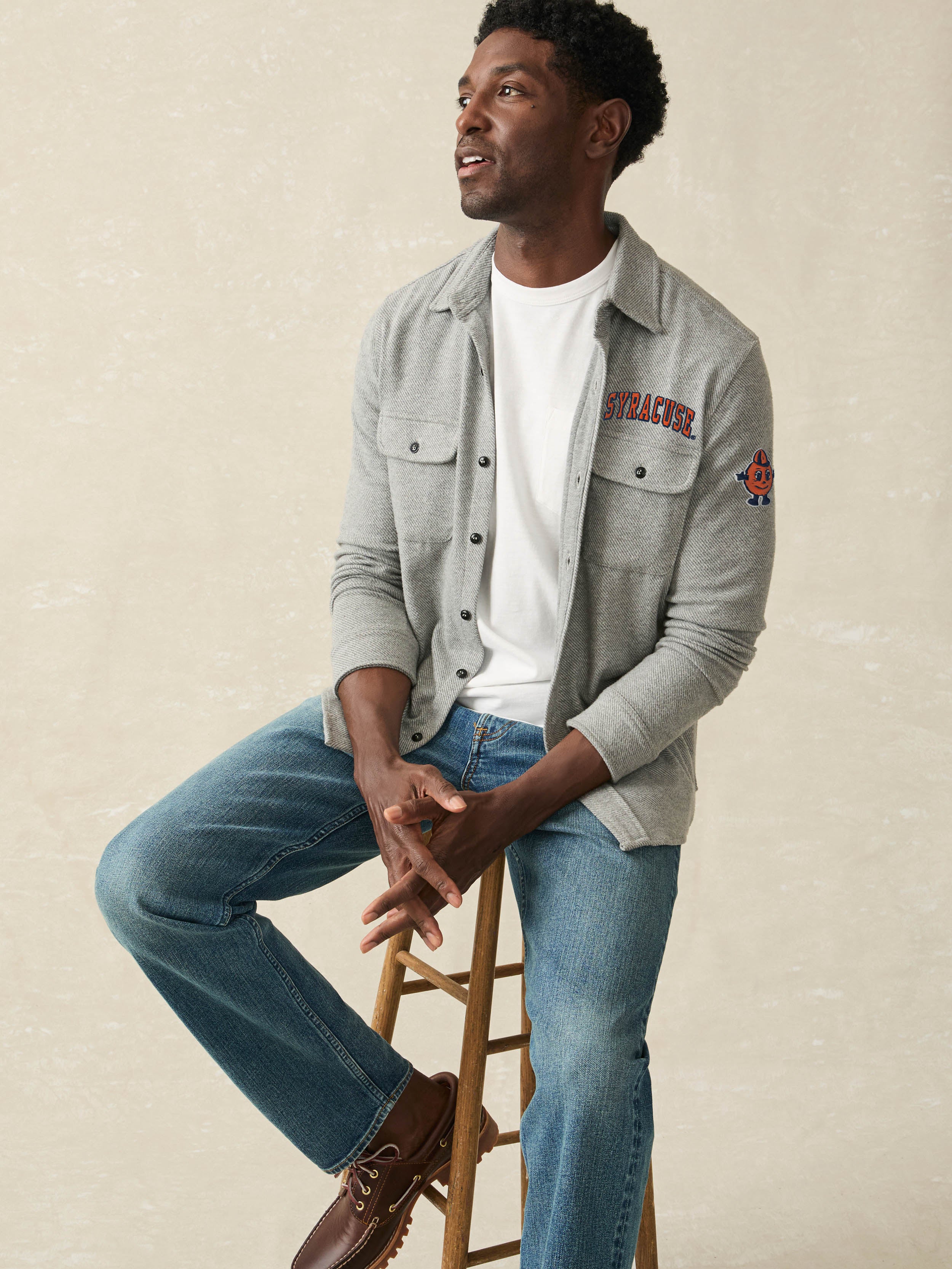 Man sitting on a stool wearing a gray long sleeve button up shirt with Syracuse University graphics at the left chest and arm with blue jeans.