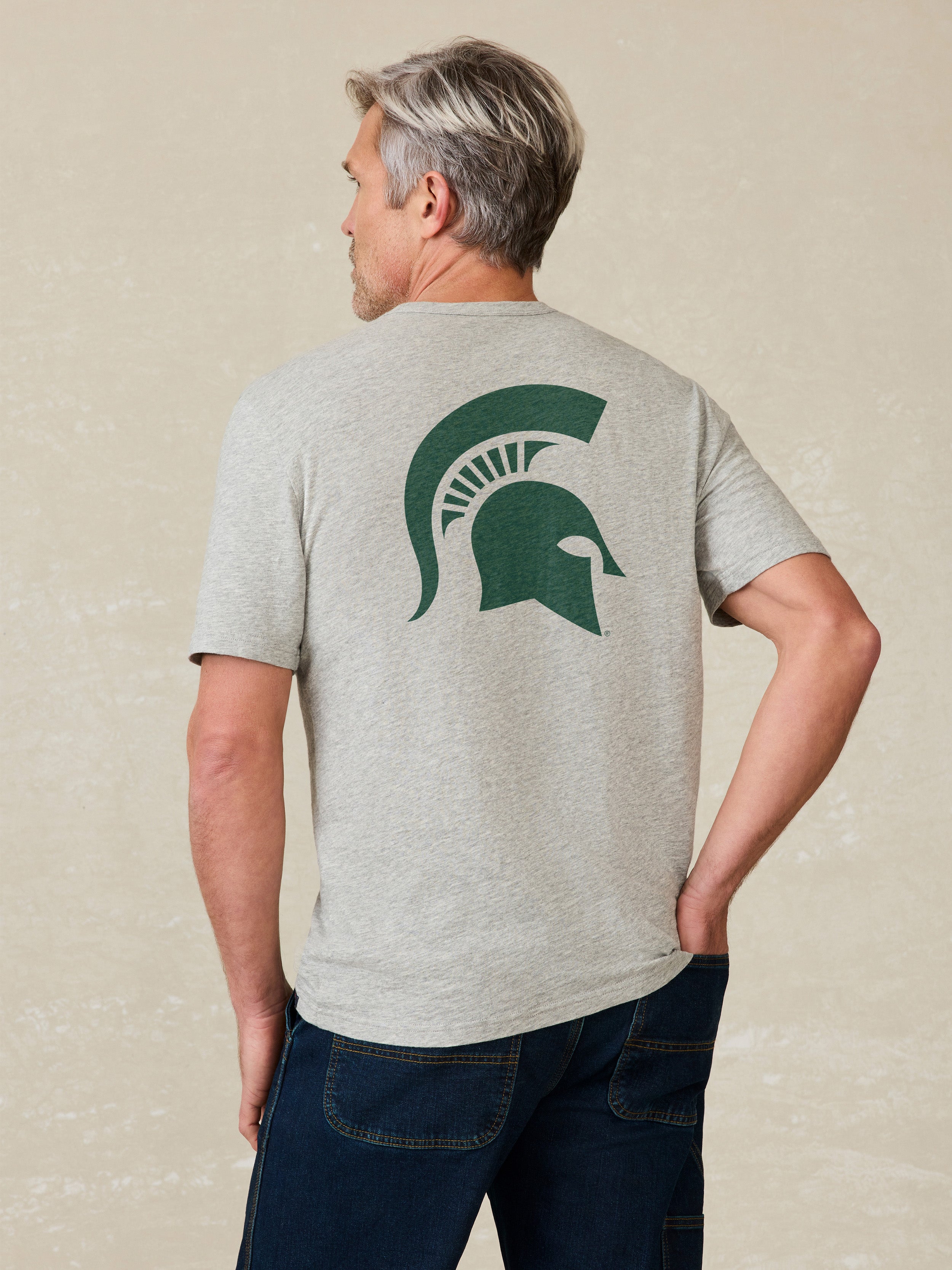 Man wearing a gray t-shirt with a green Michigan State graphic logo on the back against a beige background