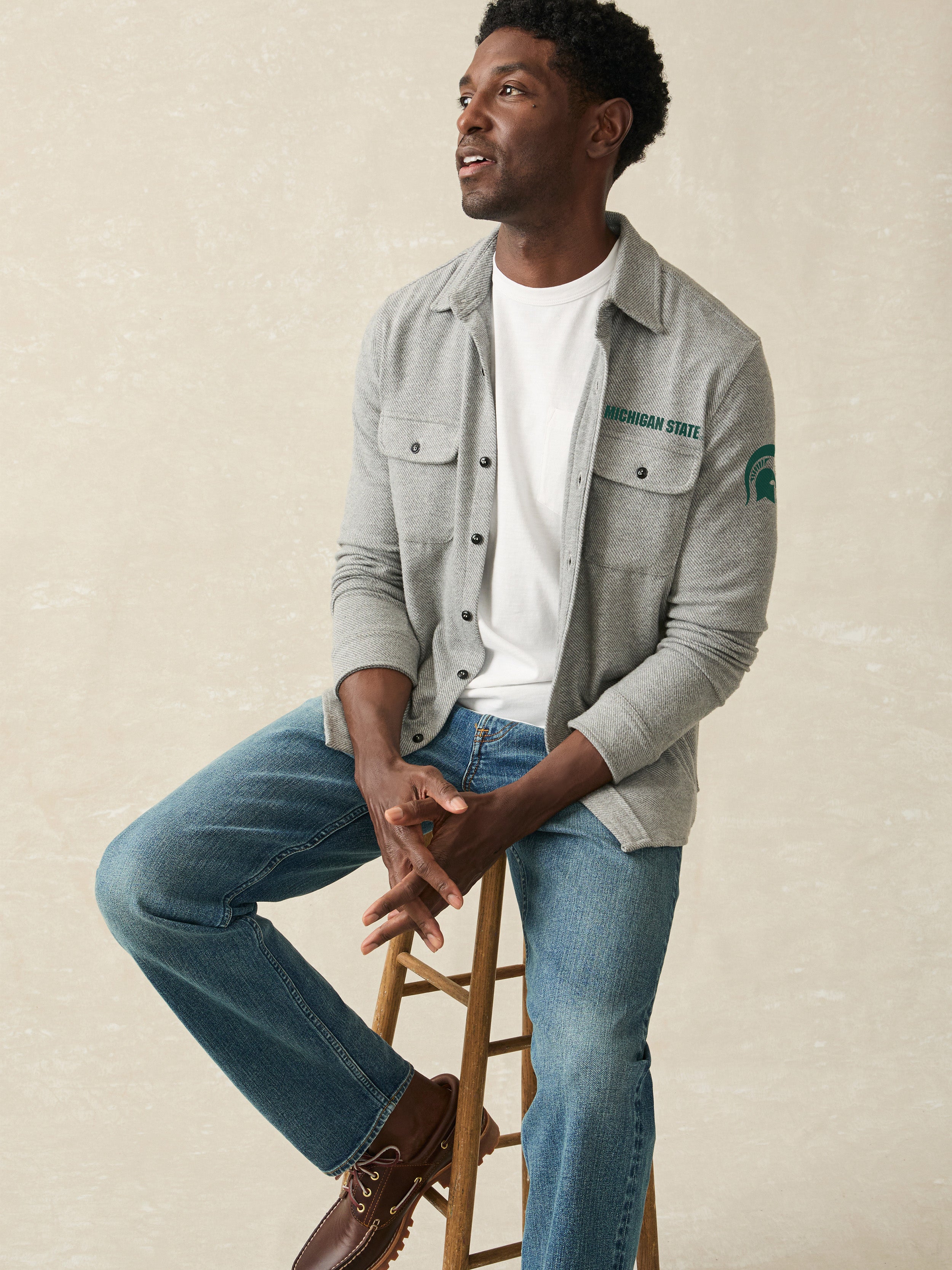 Man sitting on a stool wearing a gray shirt with a Michigan State logo at the left arm, white t-shirt, and blue jeans.