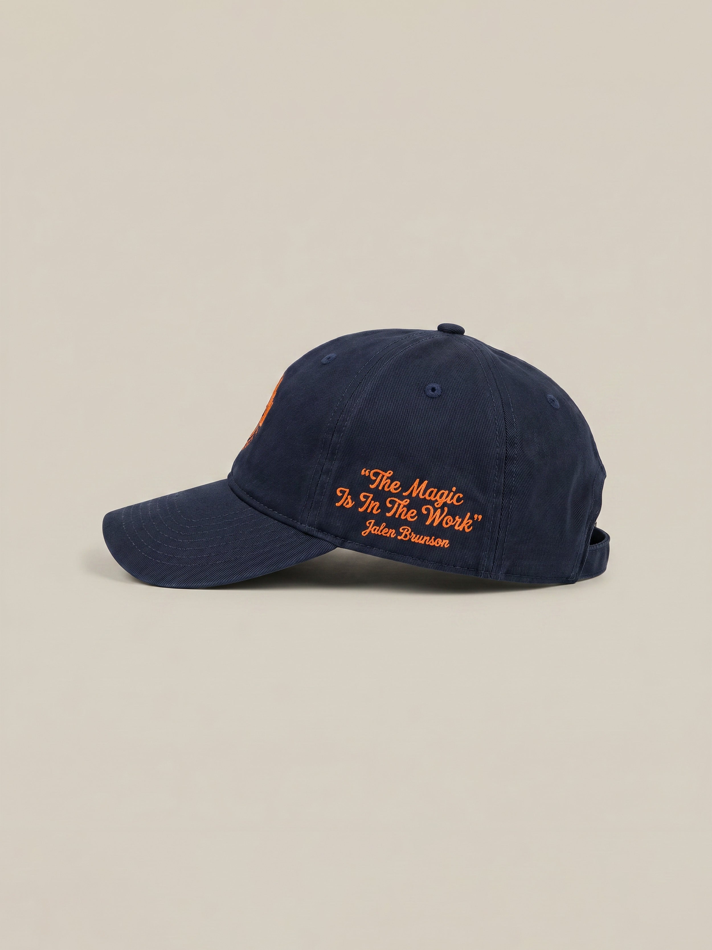 Navy blue cap with embroidered "The Magic is in the Work" text against a beige background