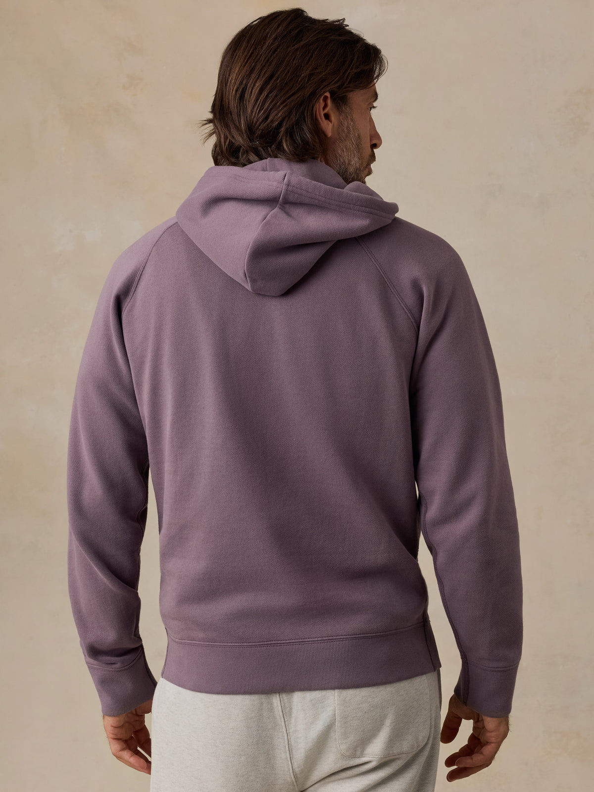 A man viewed from the back wearing a dusty purple pullover hoodie and light gray sweatpants, standing against a textured beige wall., image 3 of 5