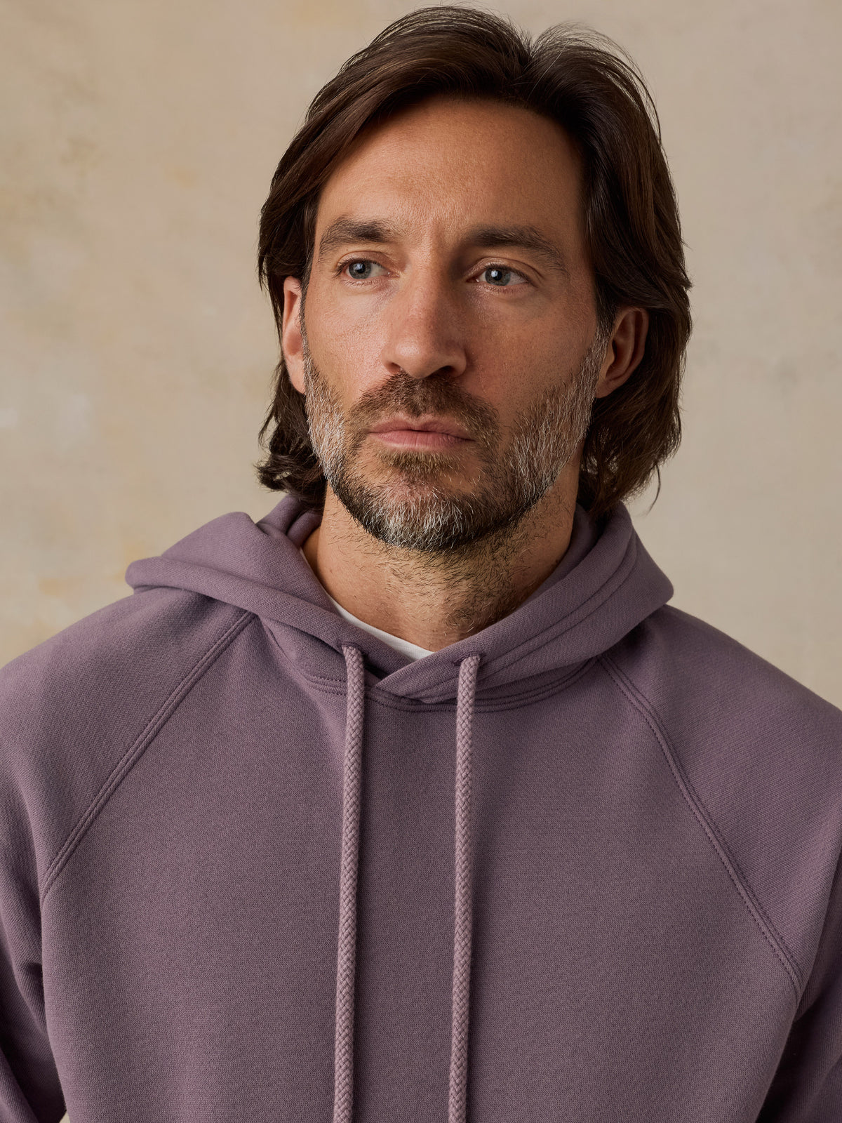 A man with brown hair and a beard wearing a dusty purple pullover hoodie, photographed from the chest up against a soft beige textured background., image 2 of 5