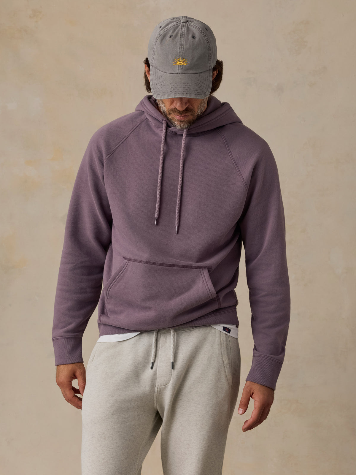 A man wearing a dusty purple pullover hoodie, light gray sweatpants, and a gray baseball cap, standing against a textured beige background., image 5 of 5
