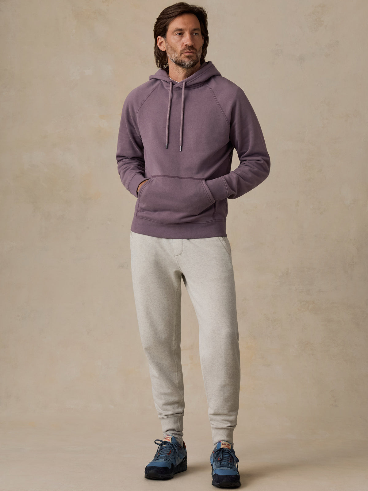 A man wearing a dusty purple pullover hoodie, light gray sweatpants, and blue sneakers, standing against a textured beige studio backdrop., image 4 of 5