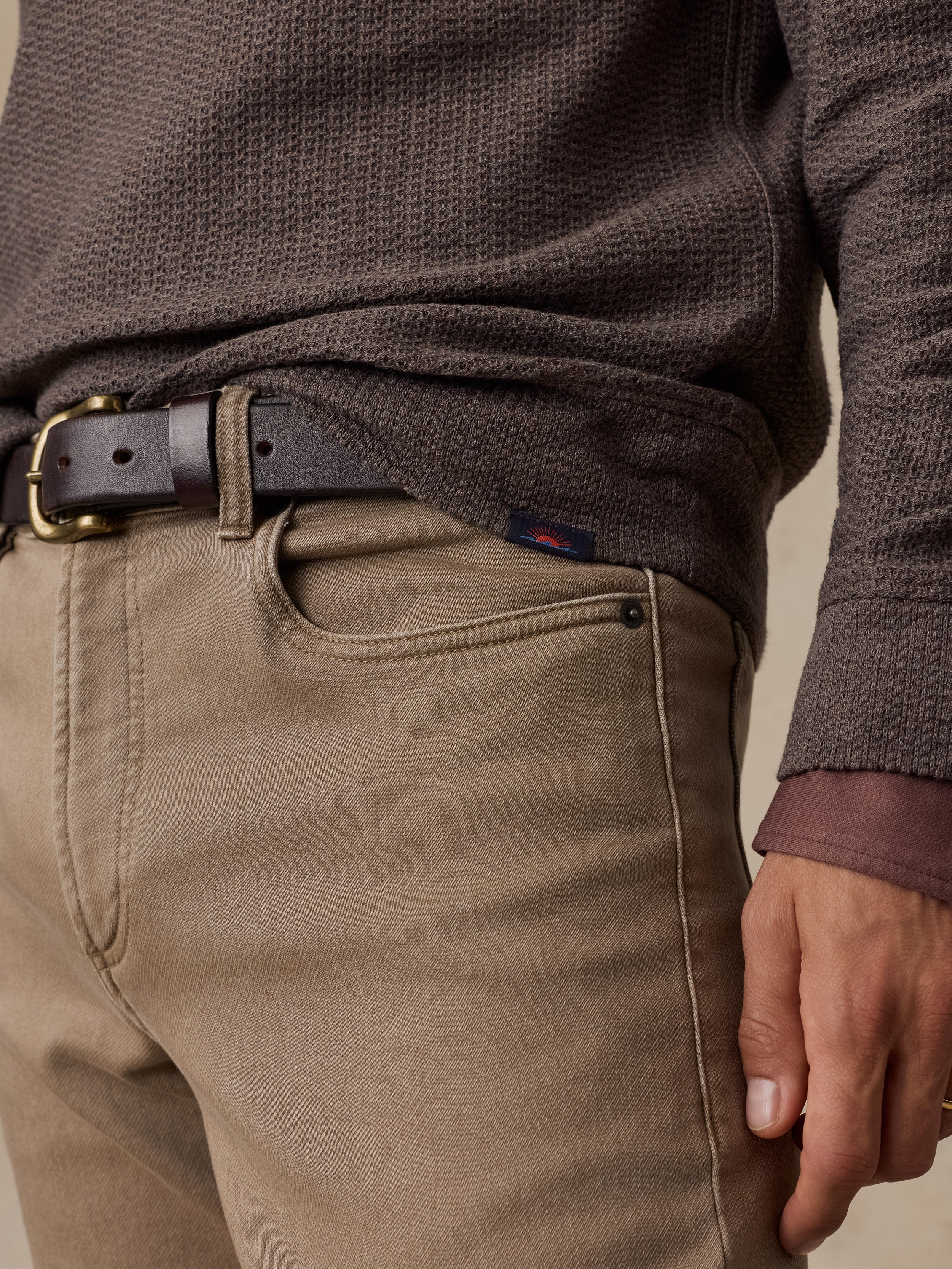 Close-up of a man wearing a dark brown textured knit sweater, a dark brown leather belt with a gold buckle, and tan trousers.
