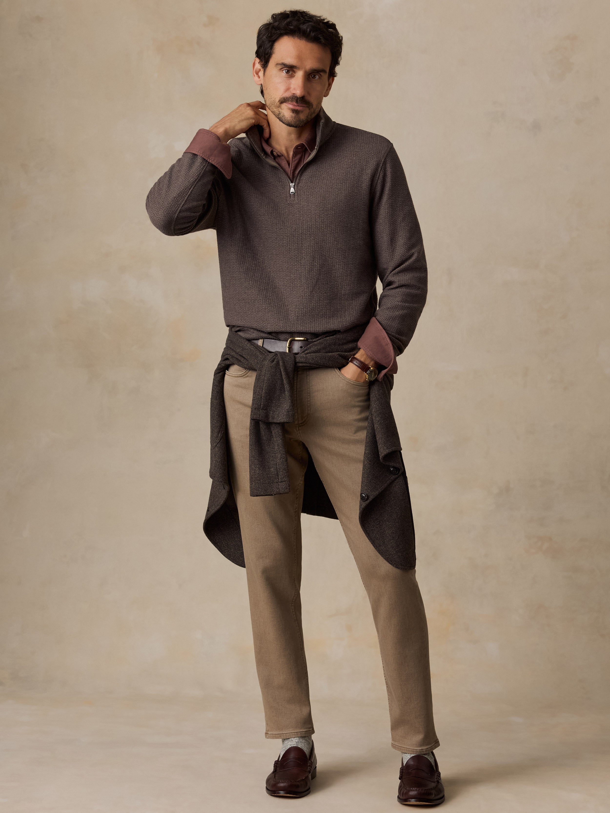 A man modeling a dark brown quarter-zip knit sweater with contrasting brown cuffs, paired with beige trousers and dark brown leather loafers, standing against a textured beige studio backdrop.