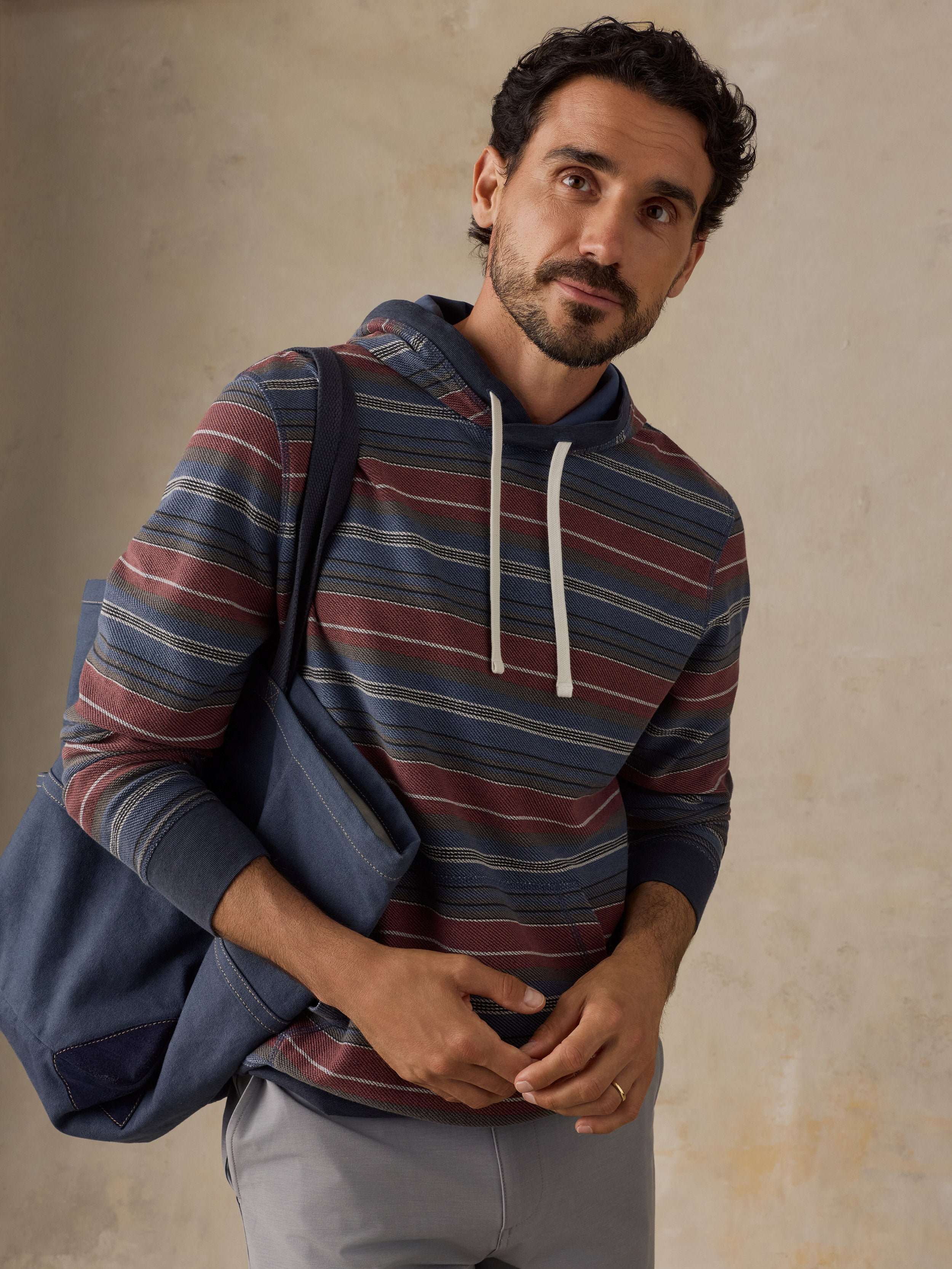 A man wearing a blue, red, and beige striped hoodie with white drawstrings, carrying a navy blue tote bag over his shoulder, set against a textured beige wall.