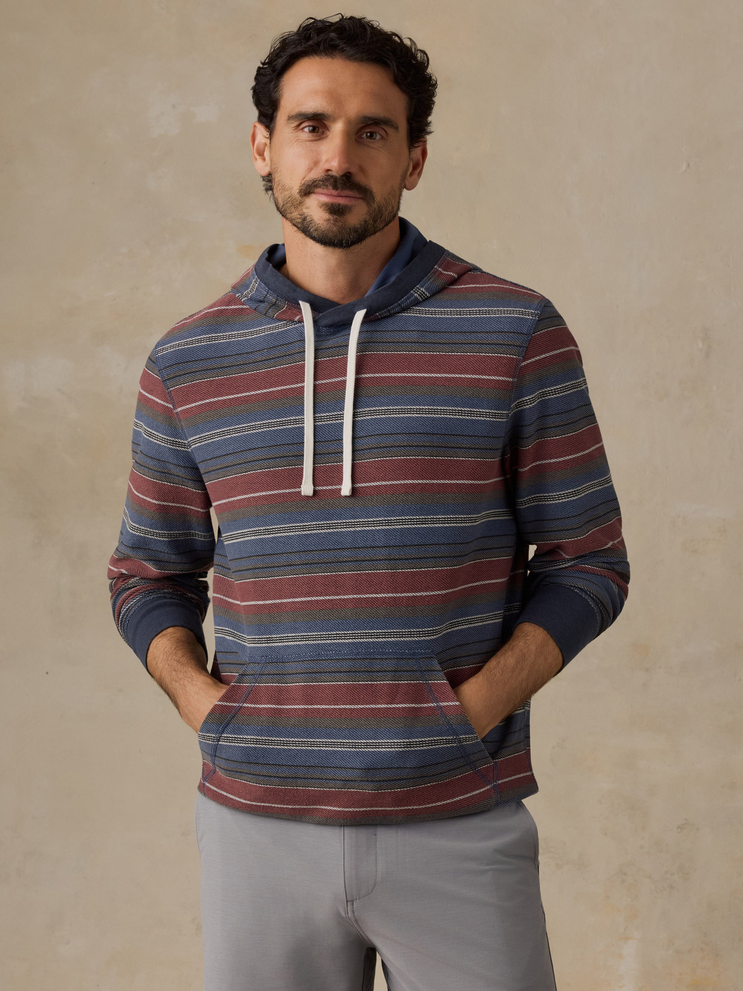 A man wearing a long-sleeve striped hoodie with blue, red, and beige horizontal stripes, paired with light gray trousers, standing against a textured beige wall.