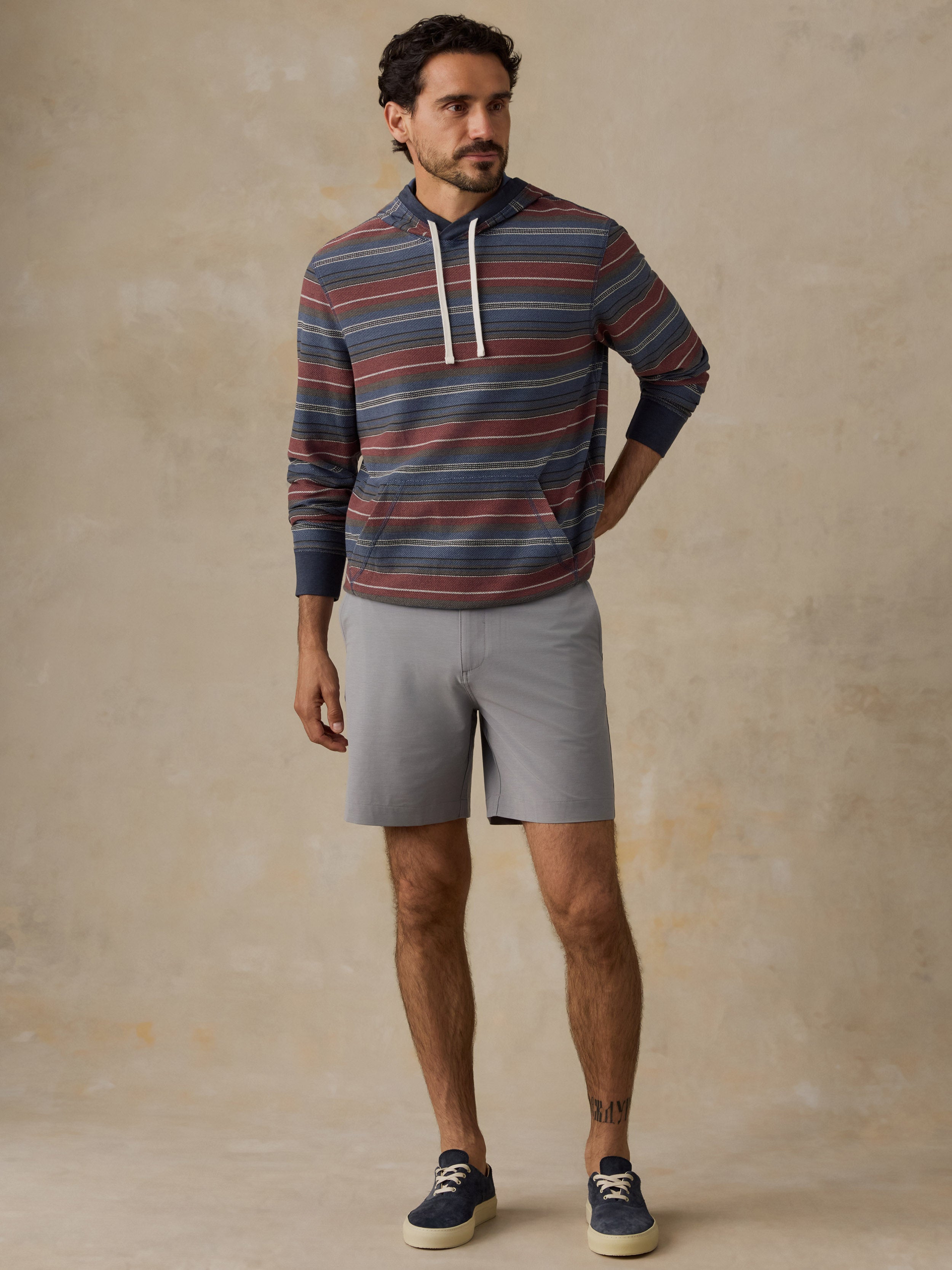 A man wearing a long-sleeve striped hoodie with navy, red, and gray horizontal stripes, white drawstrings, gray shorts, and navy blue sneakers, standing against a textured beige studio backdrop.
