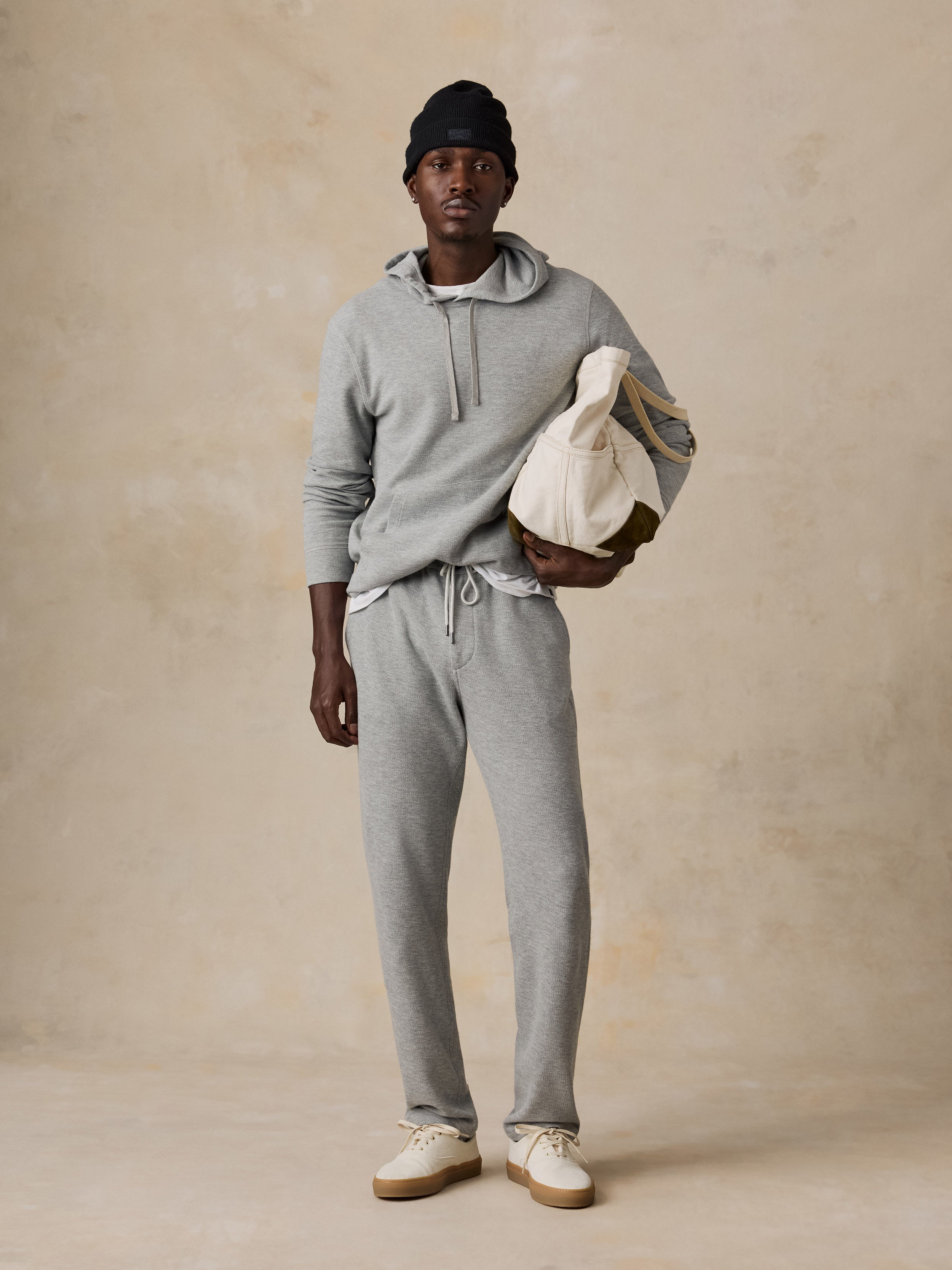A man modeling a light gray heathered hoodie and matching sweatpants, holding a cream canvas tote bag, set against a textured beige studio backdrop.