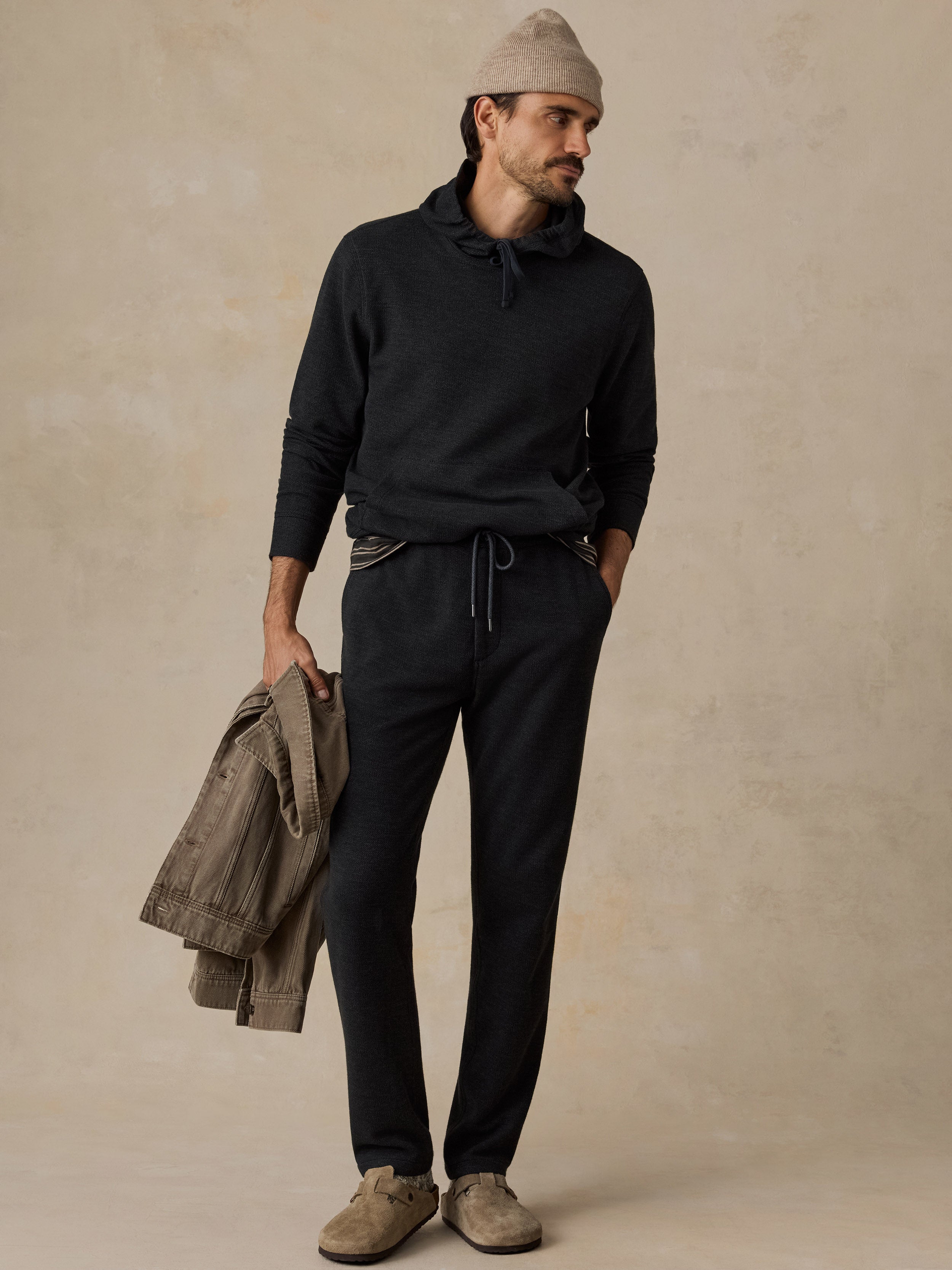 A man stands against a textured beige studio backdrop wearing a dark charcoal hoodie, matching dark drawstring sweatpants, a beige knit beanie, and taupe suede shoes, while holding a light brown jacket.