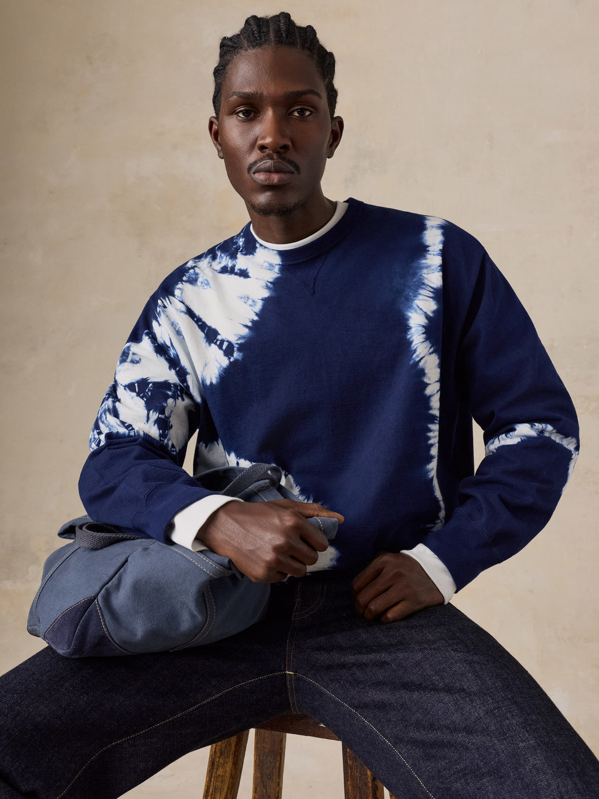 A man sitting on a stool wearing a shibori dyed sweatshirt and blue jeans, image 5 of 6