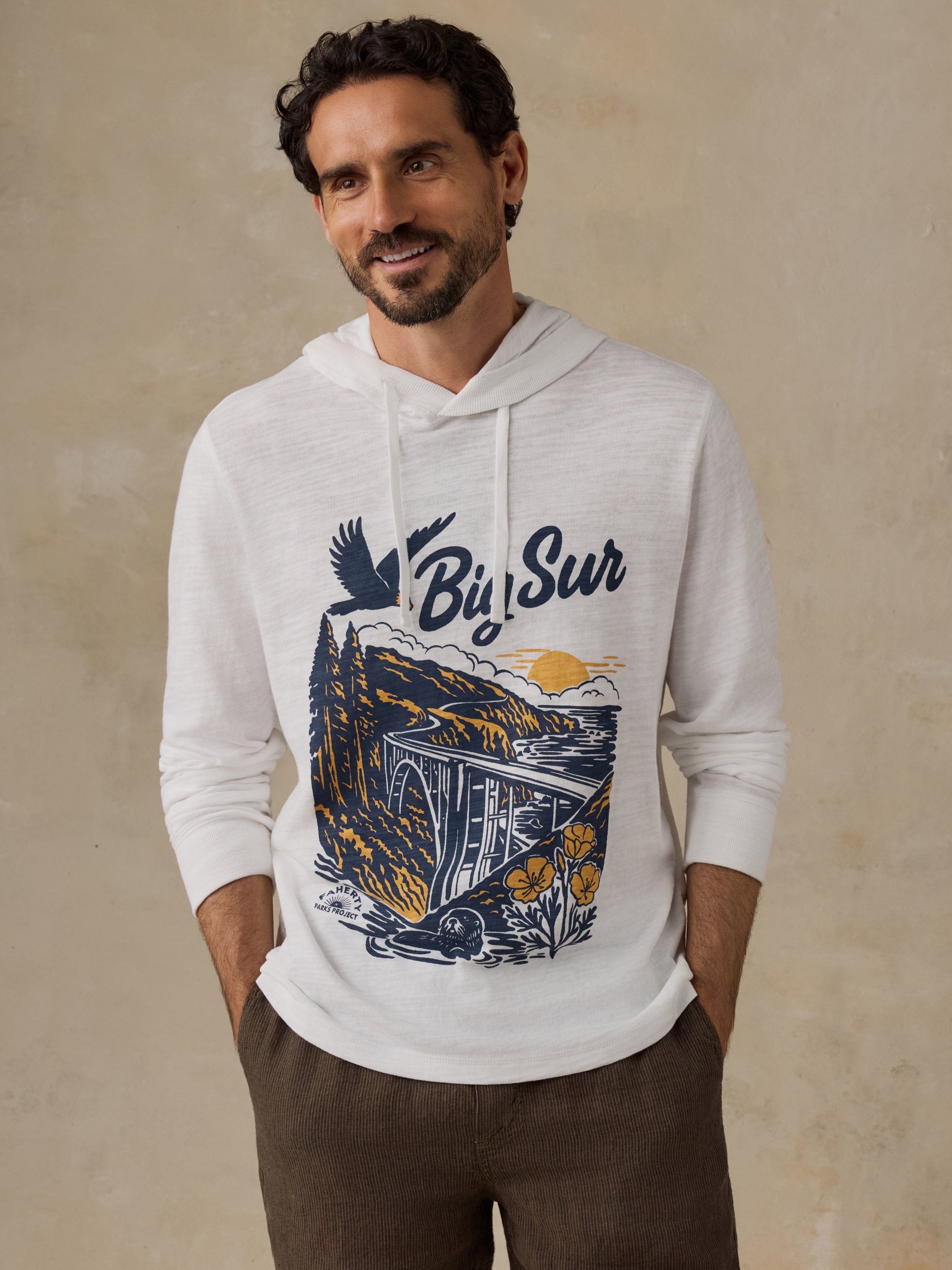 A man wearing a white long-sleeve hoodie with a graphic print of a van, mountains, and the words 'Big Sur' in navy blue and yellow, paired with brown pants, standing against a textured beige wall.