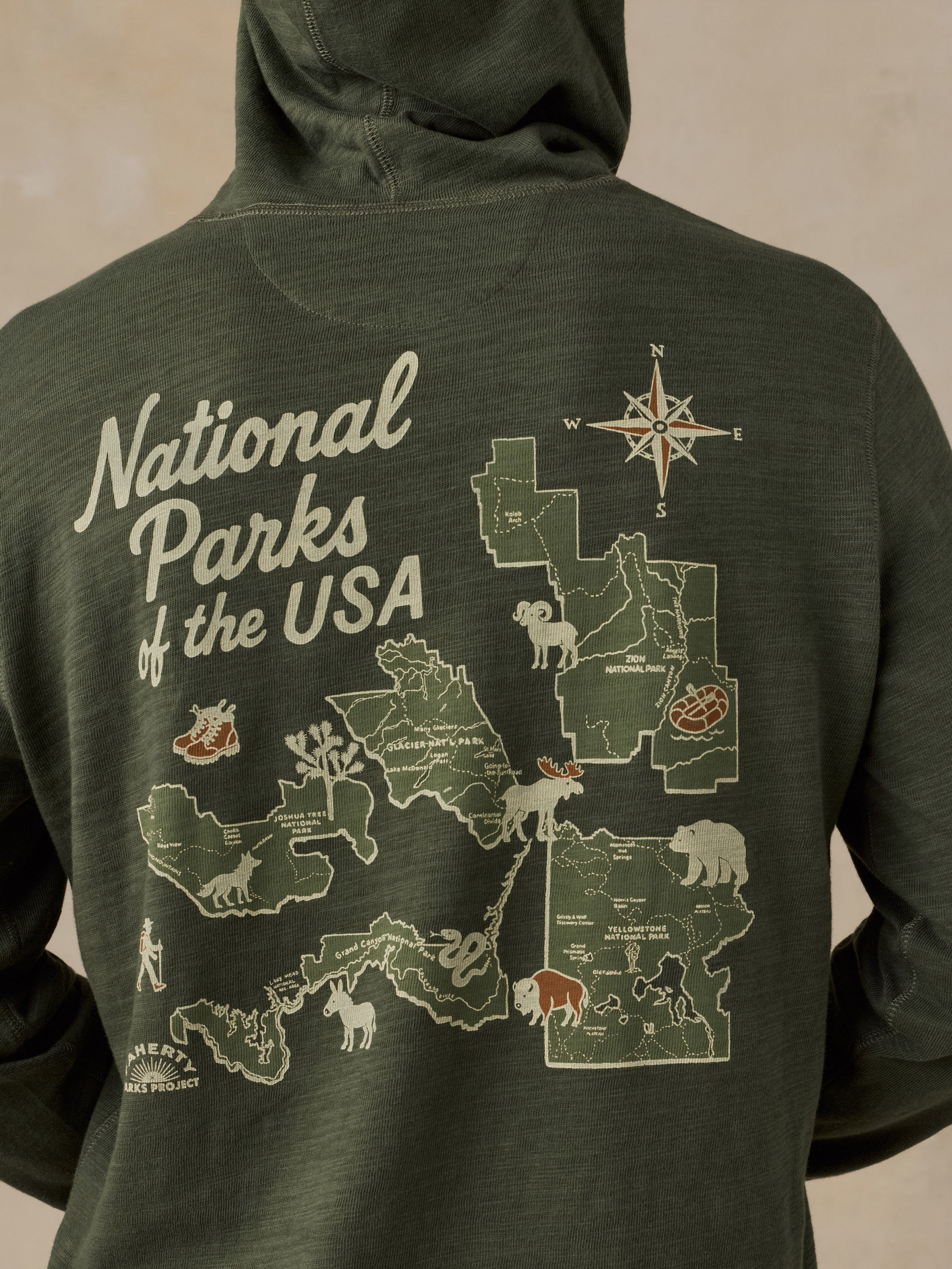 A close-up of the back of a dark olive green hooded sweatshirt featuring a large graphic print of National Parks of the USA, including state outlines, wildlife illustrations, and a compass rose.