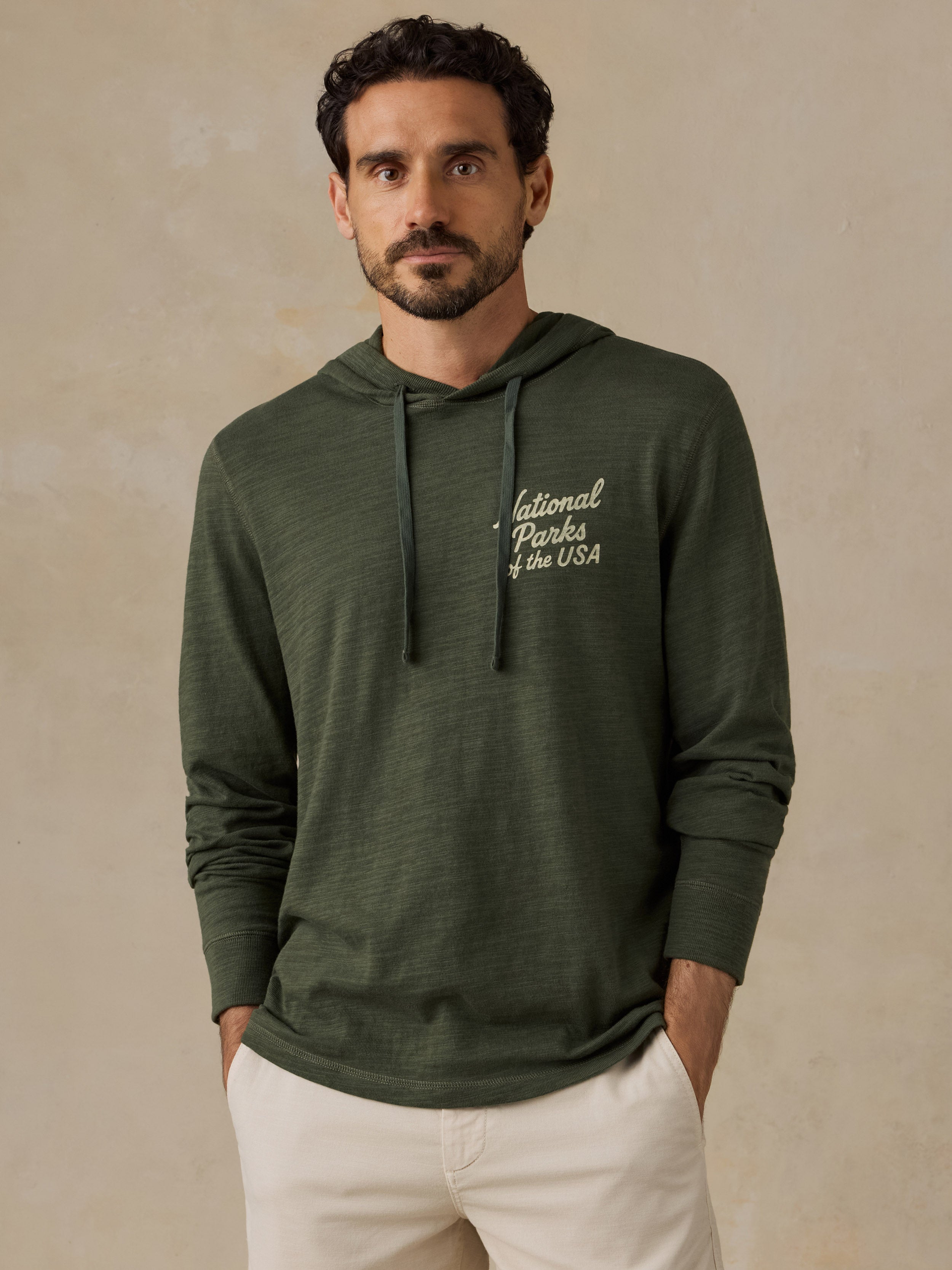 A man wearing an olive green long-sleeve hoodie with 'National Parks of the USA' printed in white script on the left chest, paired with off-white shorts, set against a textured beige background.
