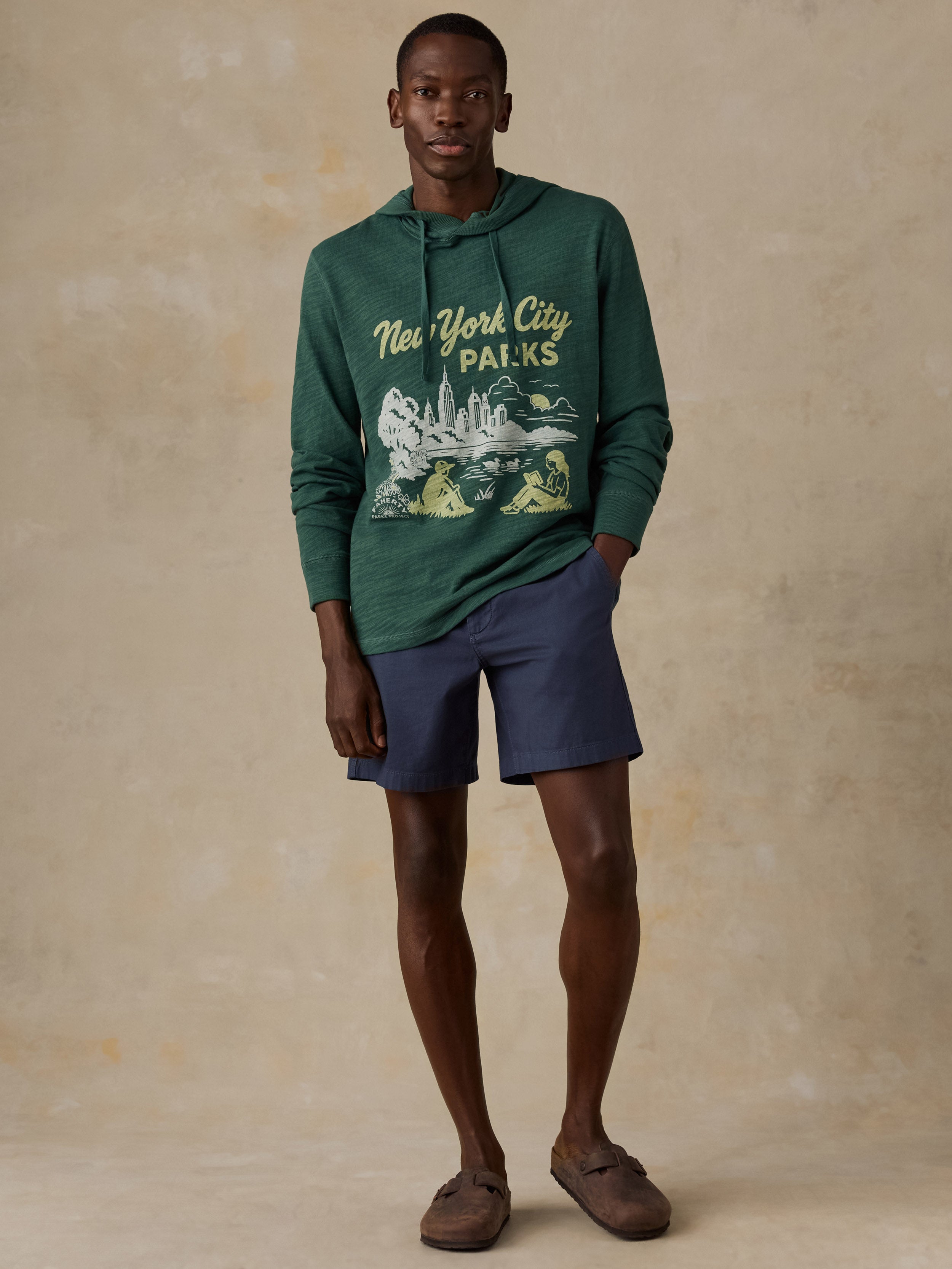 A man modeling a dark green pullover hoodie with a 'New York City Parks' graphic on the front, paired with navy blue shorts and brown slip-on shoes, against a textured beige studio backdrop.
