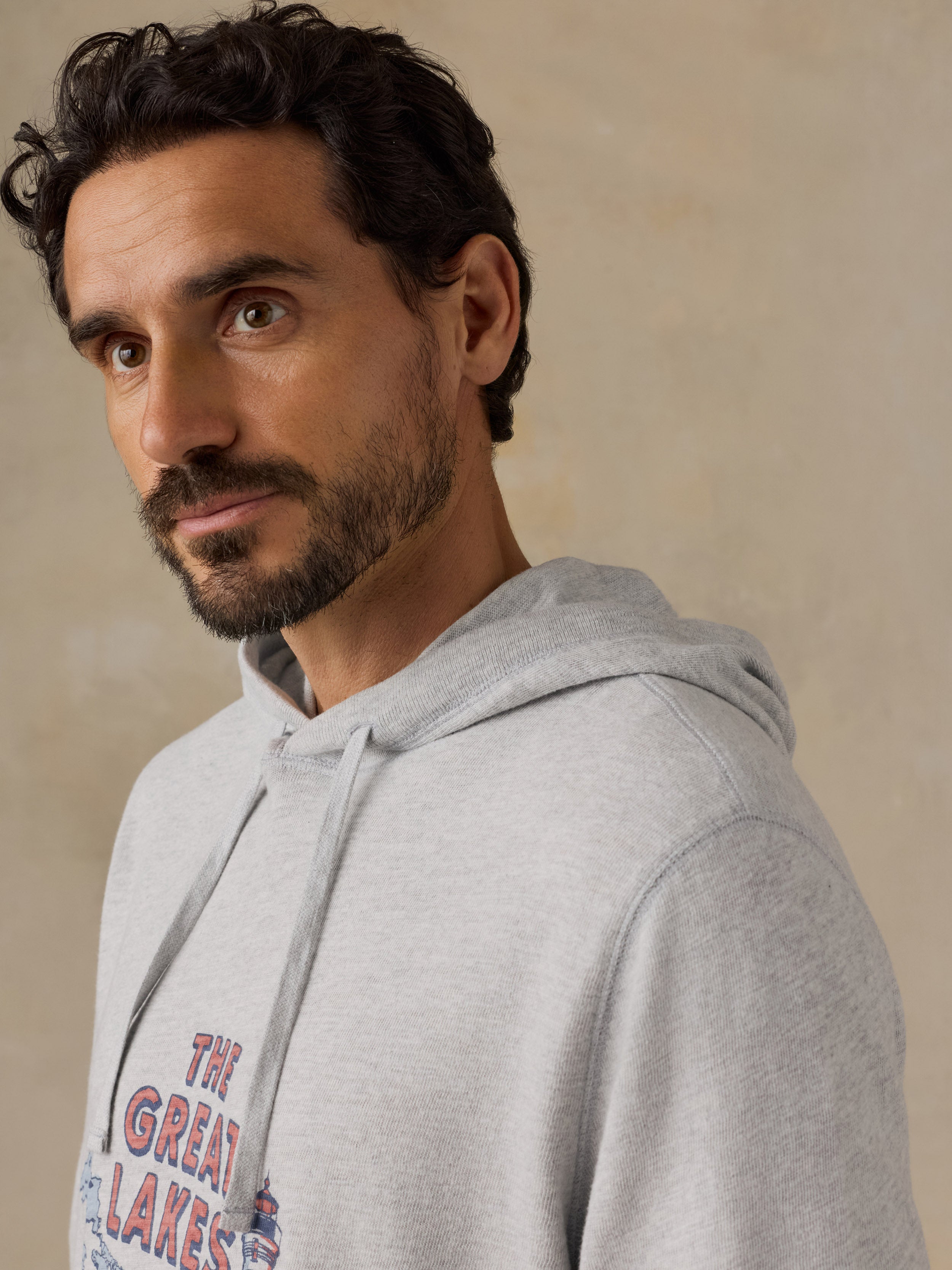 A man wearing a light gray heather hoodie with a graphic print that reads 'THE GREAT LAKES' in red and blue, photographed against a neutral beige background.