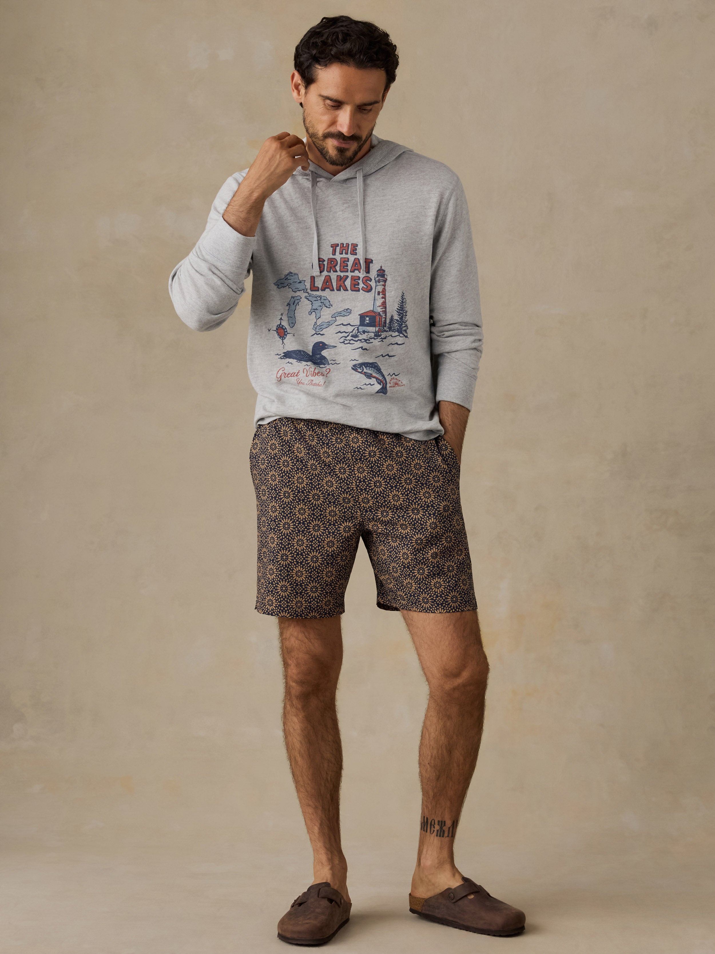 A man wearing a light gray graphic hoodie and brown patterned shorts with brown slip-on shoes, standing against a textured beige studio backdrop.
