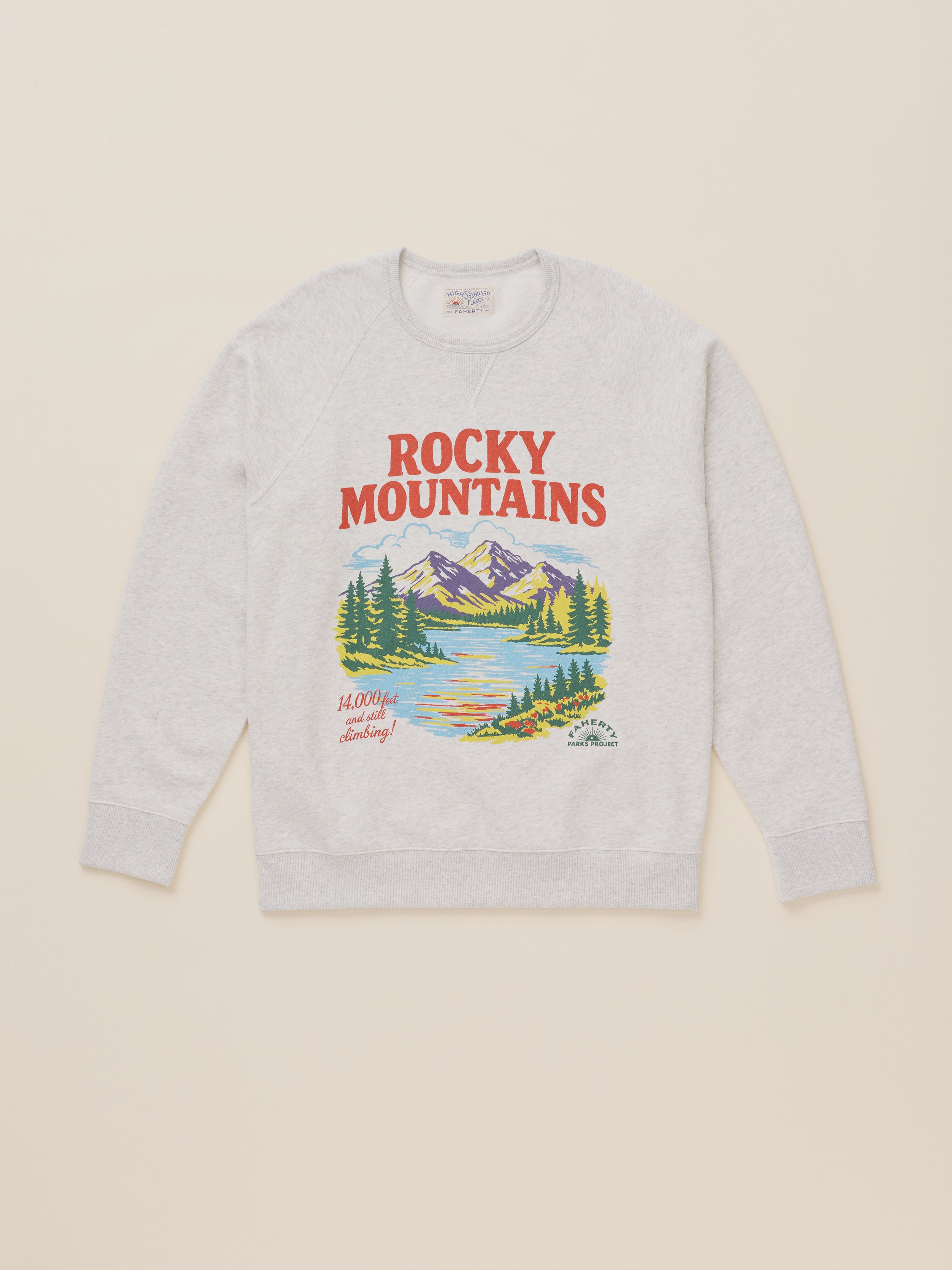 A light heather gray crewneck sweatshirt featuring a colorful graphic print of the Rocky Mountains, lakes, and pine trees, with 'ROCKY MOUNTAINS' printed in red above the illustration, laid flat against a light beige background.
