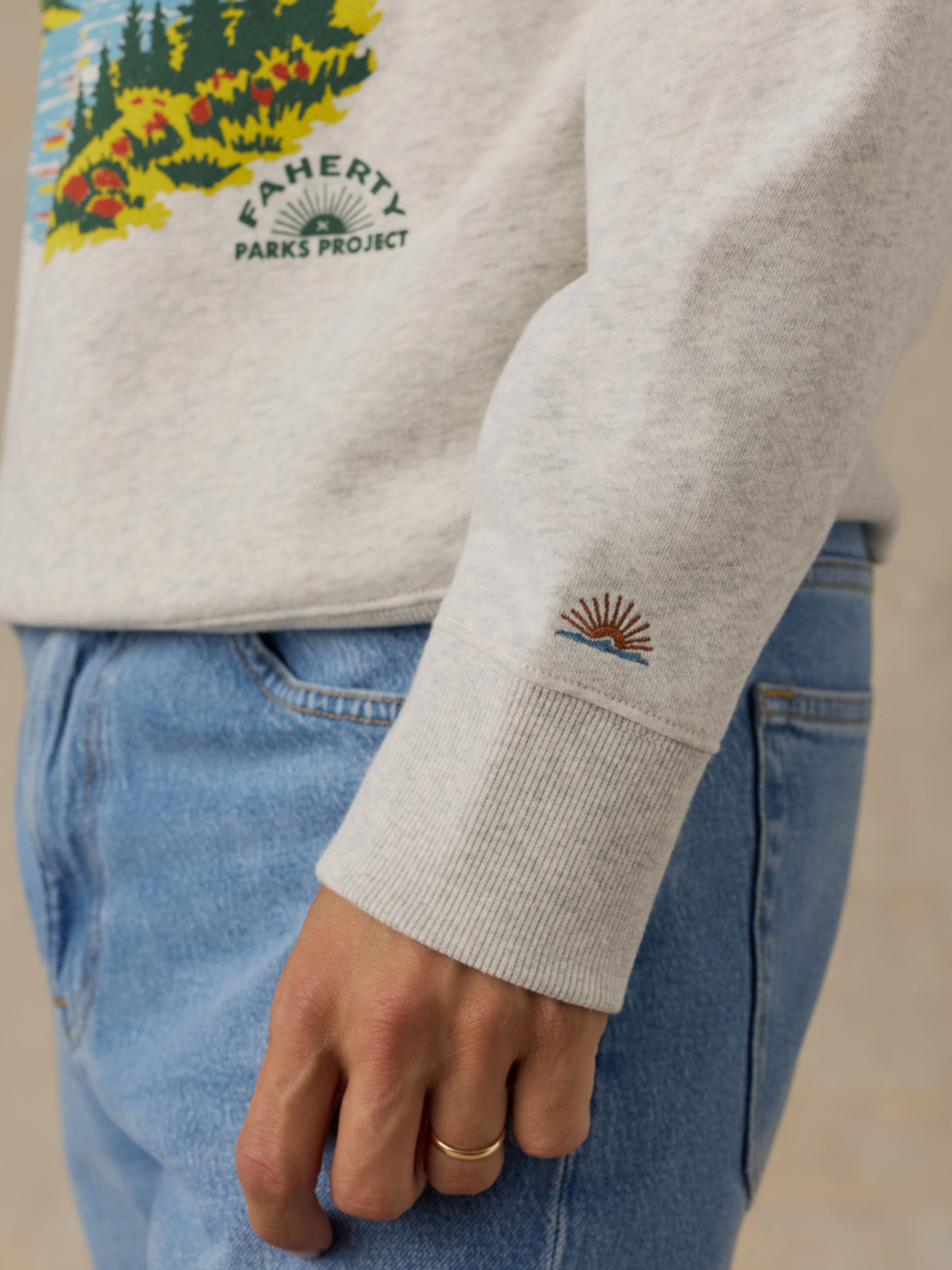Close-up of a man wearing a light heather gray sweatshirt with a colorful forest graphic and 'FAHERTY PARKS PROJECT' text on the chest, paired with light blue jeans. The sleeve has a small embroidered sunburst logo.