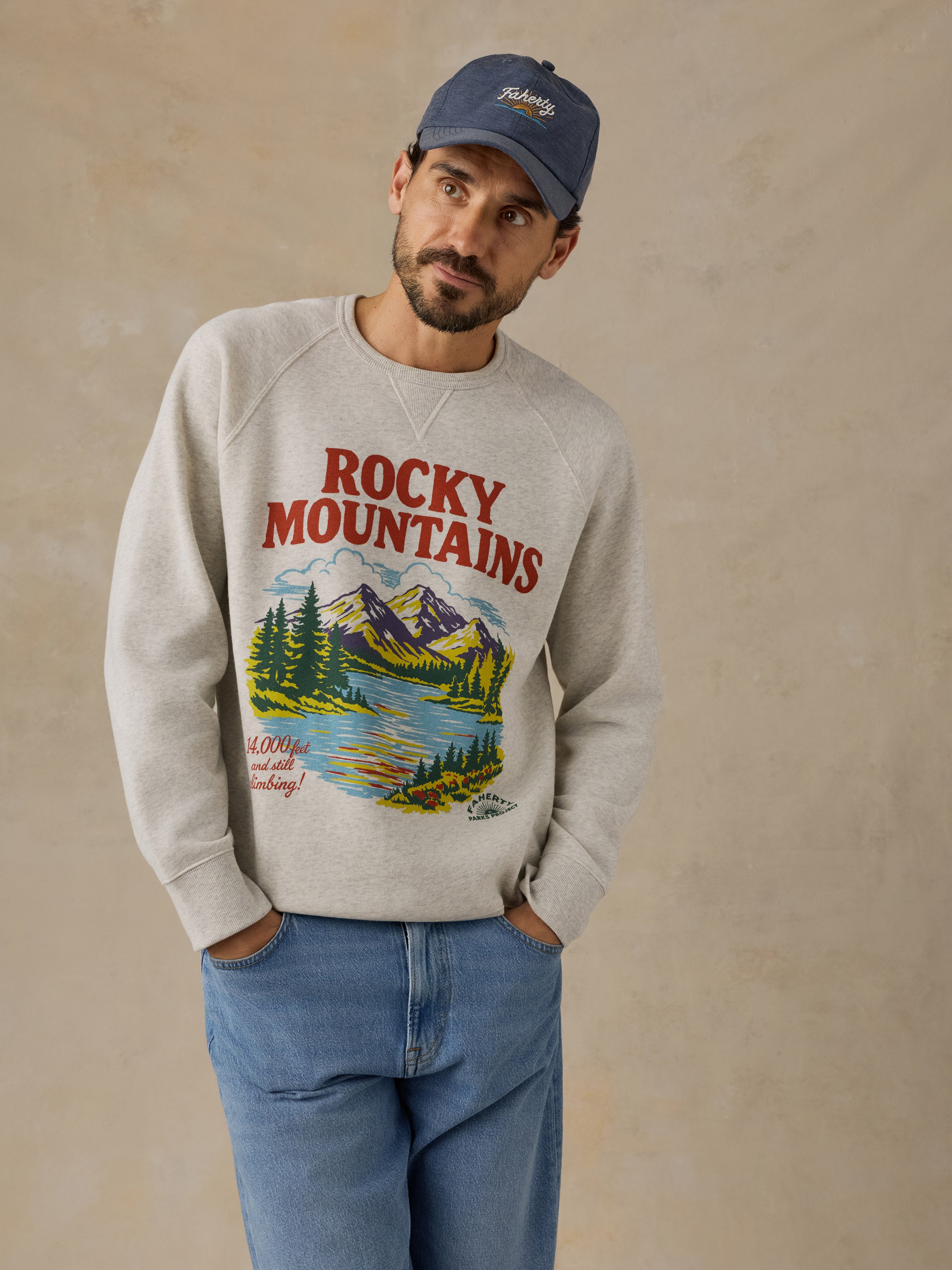 A man wearing a light gray crewneck sweatshirt with a colorful 'Rocky Mountains' graphic print on the front, paired with blue jeans and a navy blue baseball cap, standing against a textured beige wall.