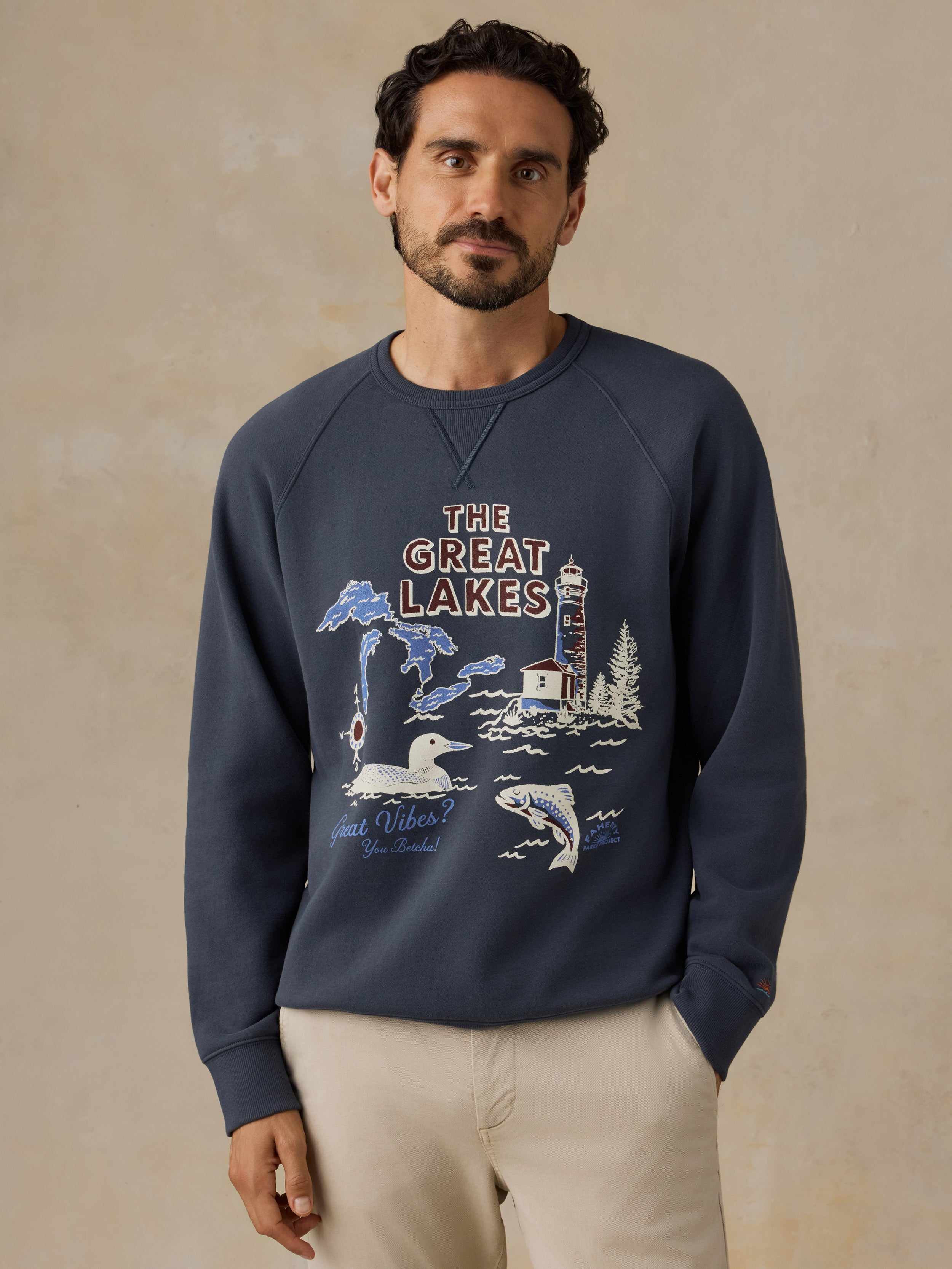 A man wearing a navy blue crewneck sweatshirt with a 'The Great Lakes' graphic print on the front, paired with beige pants, standing against a textured beige wall.