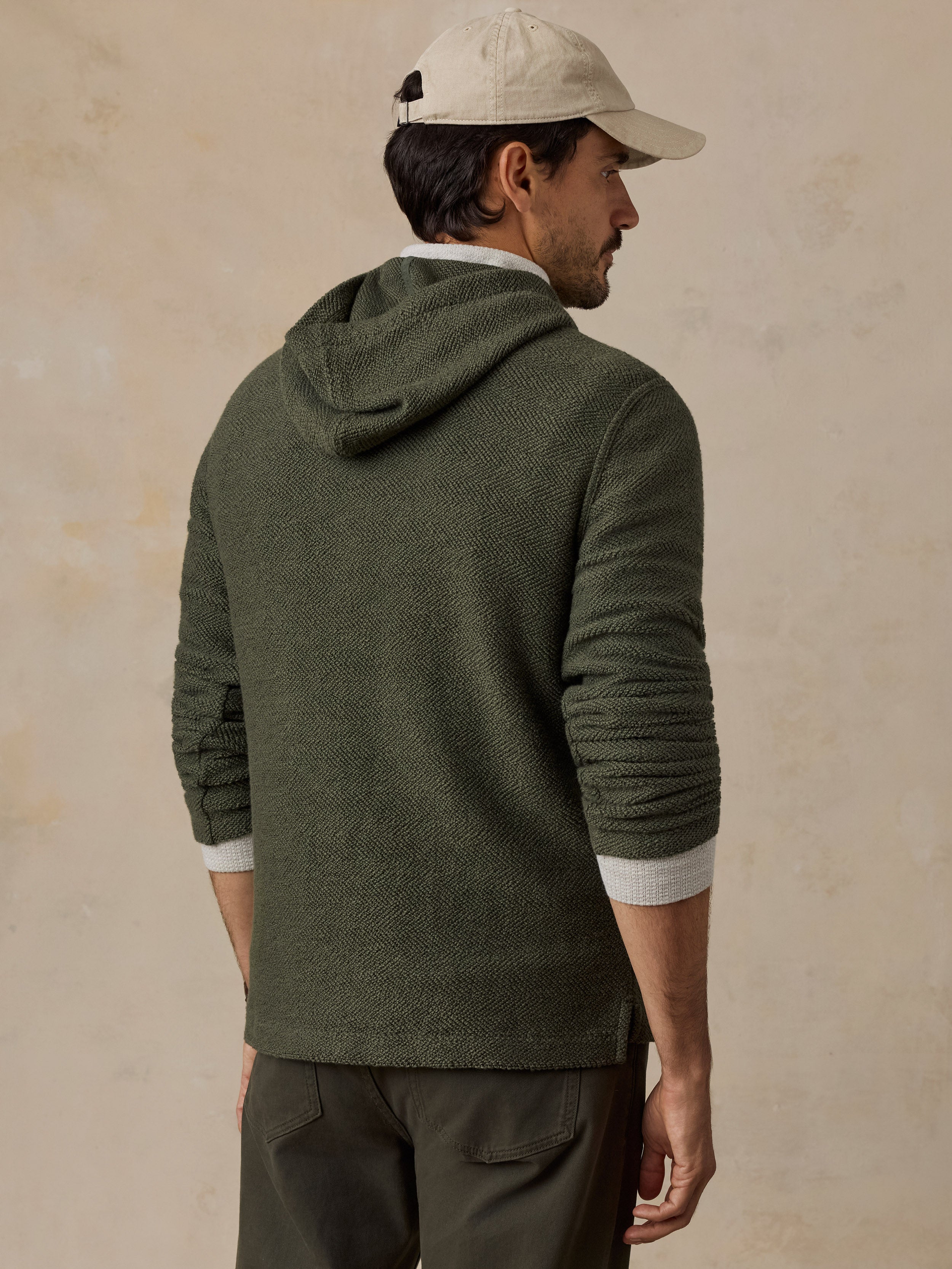 A man wearing an olive green textured knit hooded sweater, shown from the back. He is wearing a beige baseball cap, a white collared shirt underneath, and dark olive pants, set against a textured beige background.