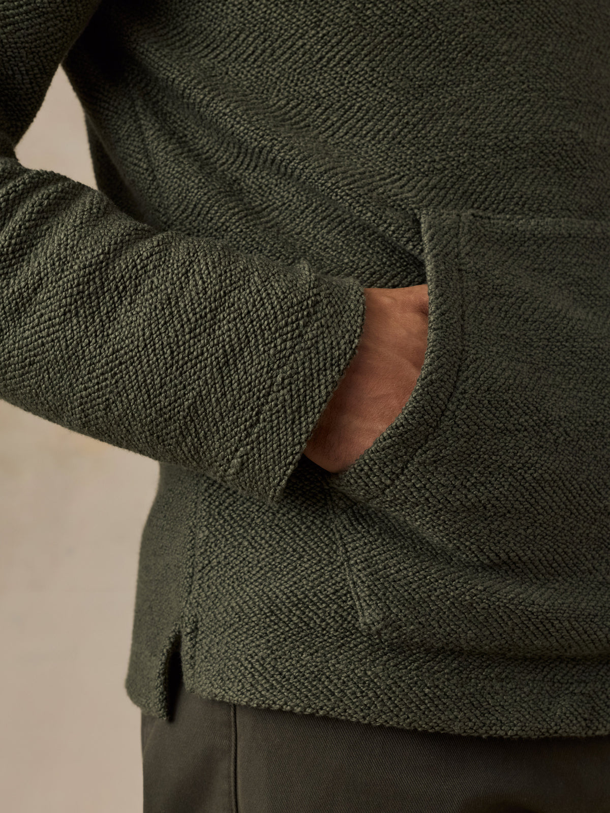 Close-up of a man wearing a dark olive green textured knit sweater with a side pocket, showing the fabric weave and a hand in the pocket.