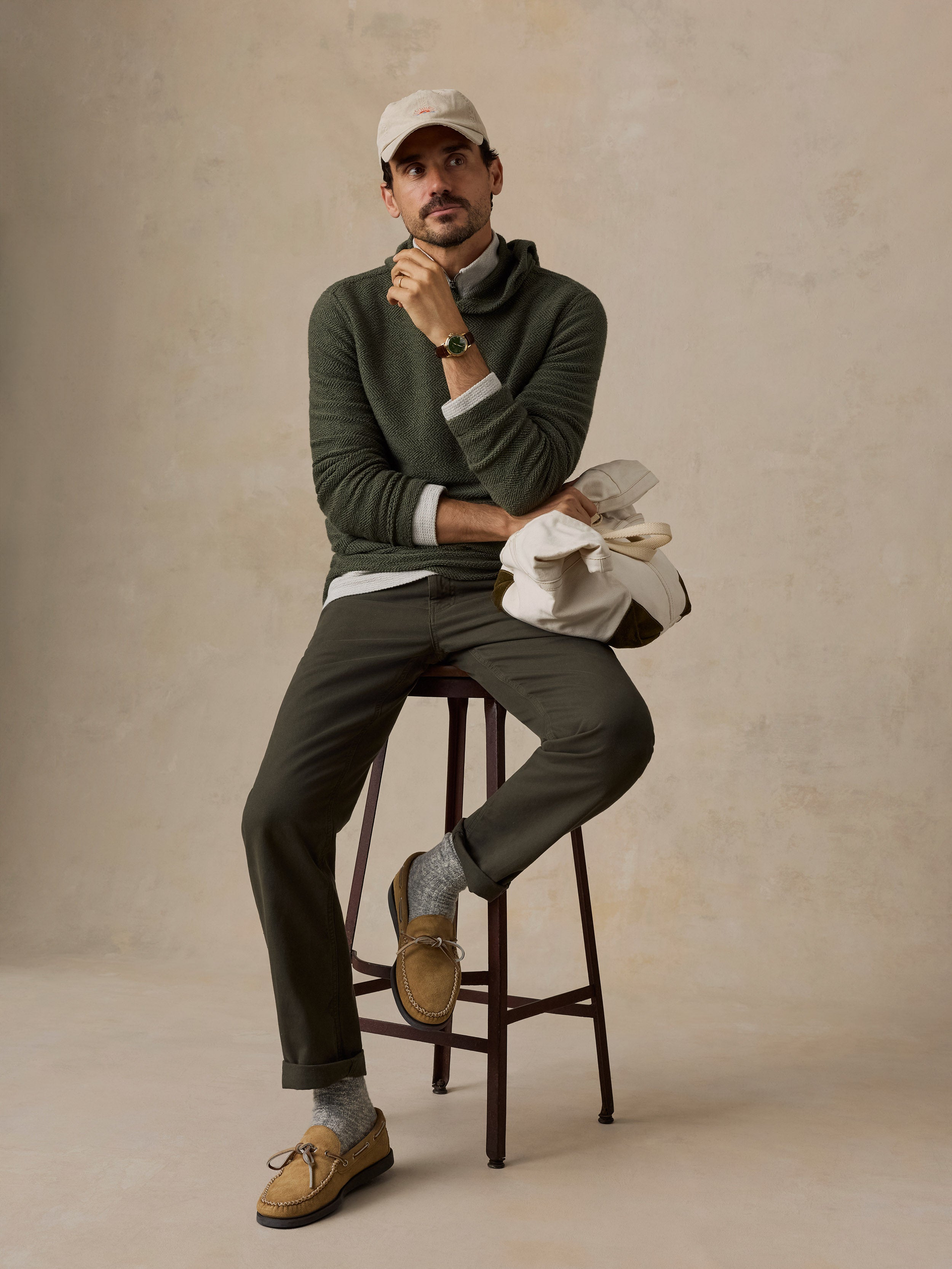 A man sits on a dark wooden stool wearing an olive green knit sweater, matching olive trousers, tan boat shoes, gray socks, and a beige baseball cap. He holds a white canvas tote bag against a textured beige studio backdrop.