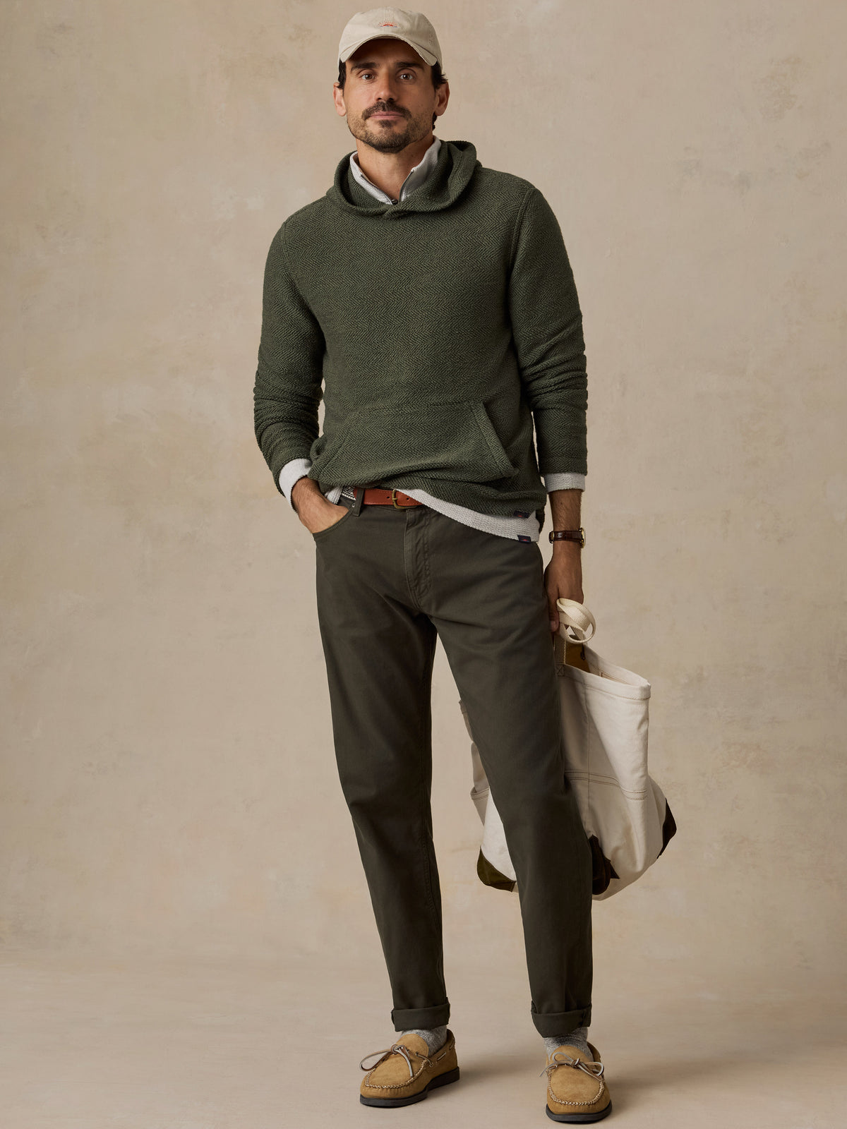 A man stands against a textured beige studio backdrop wearing an olive green knit pullover over a light gray shirt, dark olive pants, tan suede loafers, a beige cap, and holding a white tote bag.