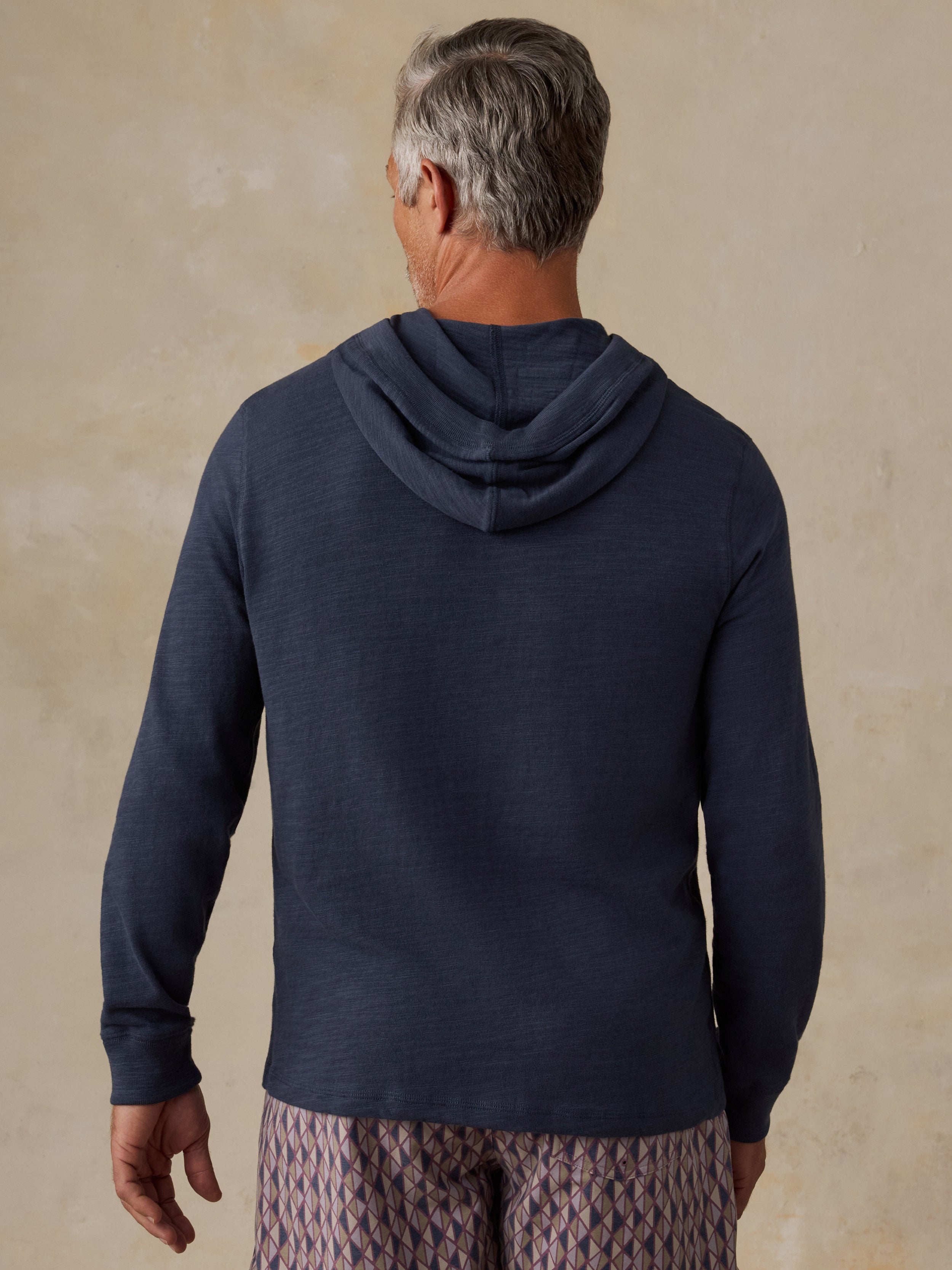 A man viewed from behind wearing a dark navy blue long-sleeve hoodie and patterned shorts, standing against a textured beige background.
