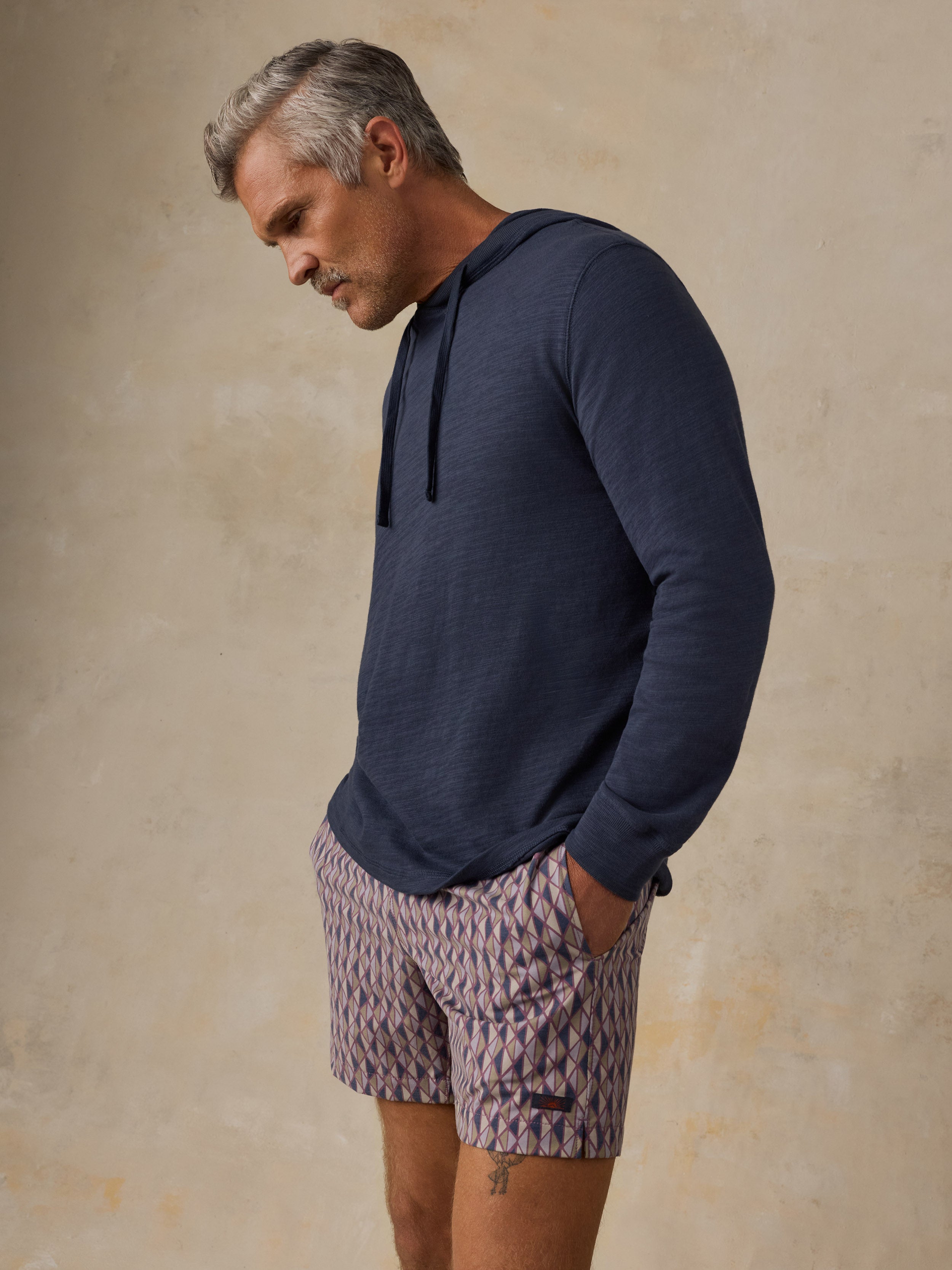 A man wearing a navy blue long-sleeve hoodie and patterned shorts, standing against a textured beige studio background.
