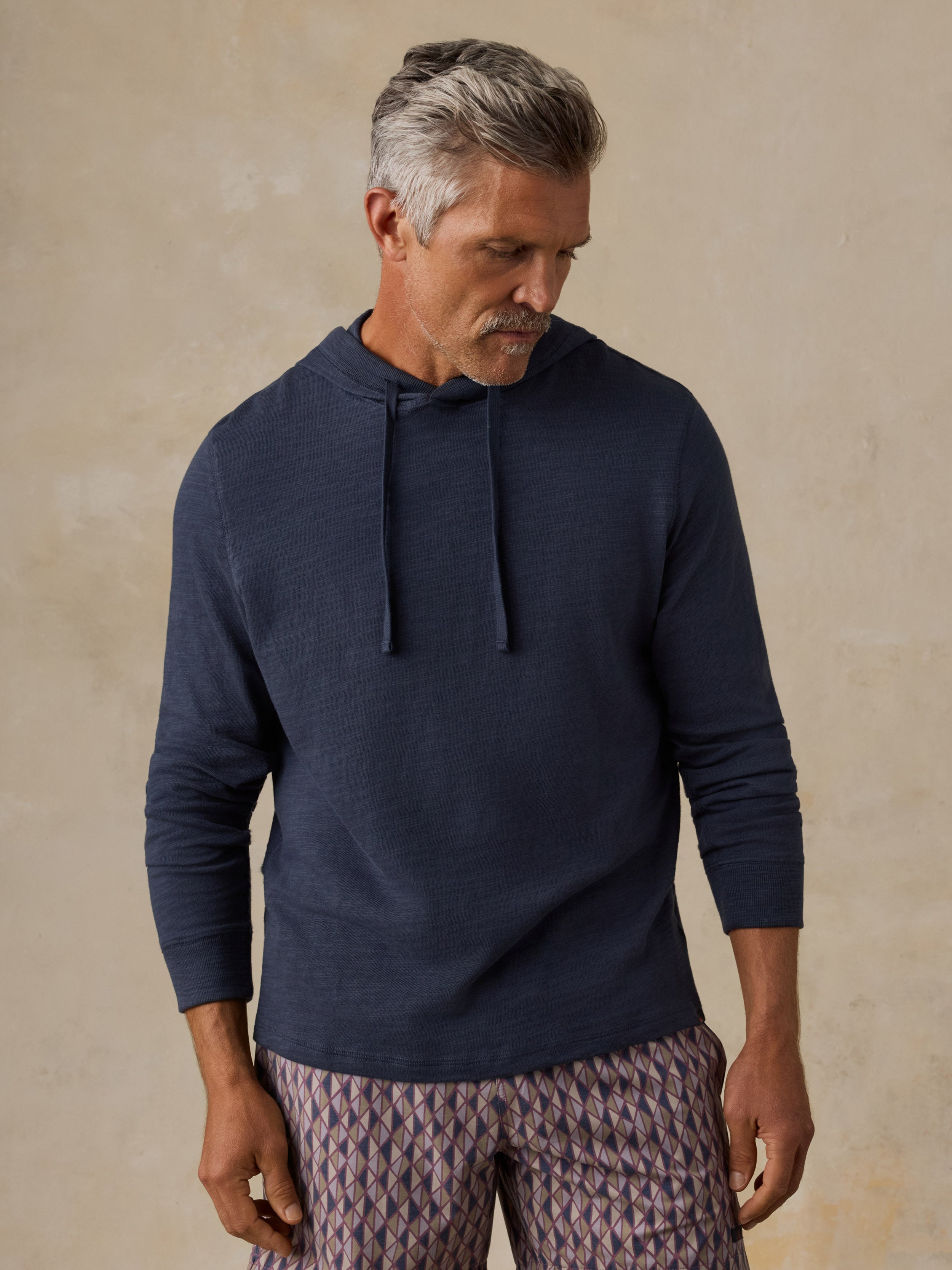 A man wearing a navy blue long-sleeve hoodie and patterned trousers, standing against a textured beige wall.