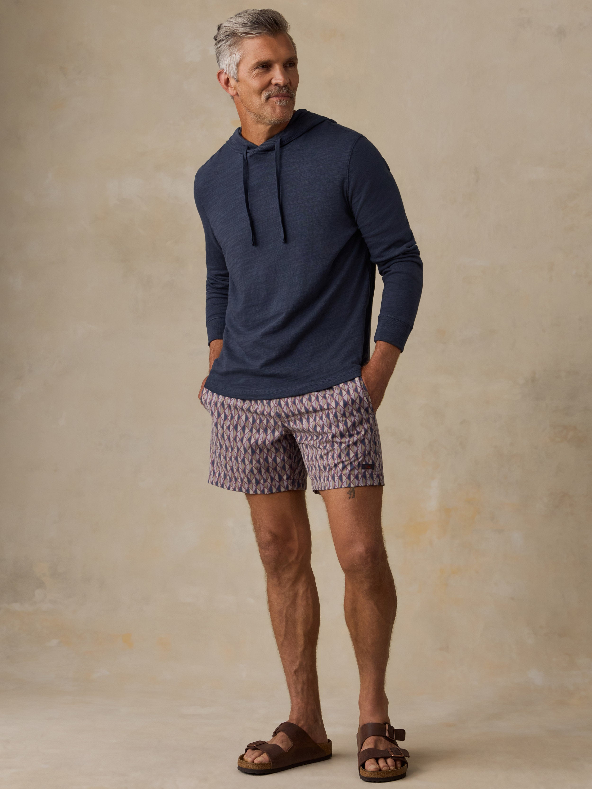 A man wearing a navy blue long-sleeve hoodie, patterned purple and white shorts, and brown sandals, standing against a textured beige studio backdrop.