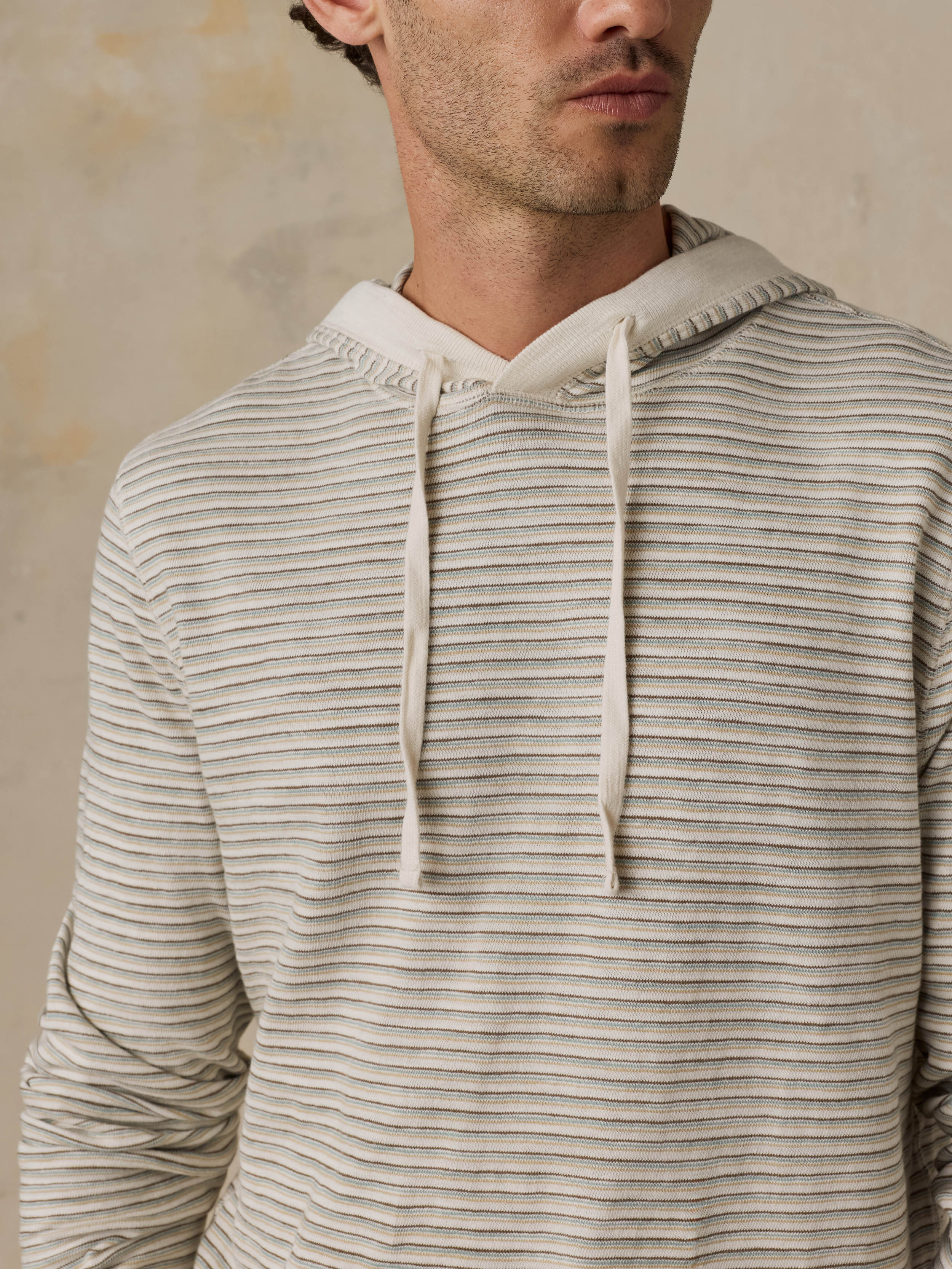 A close-up of a man wearing a light beige and off-white horizontally striped hoodie with matching drawstrings, set against a textured neutral background.