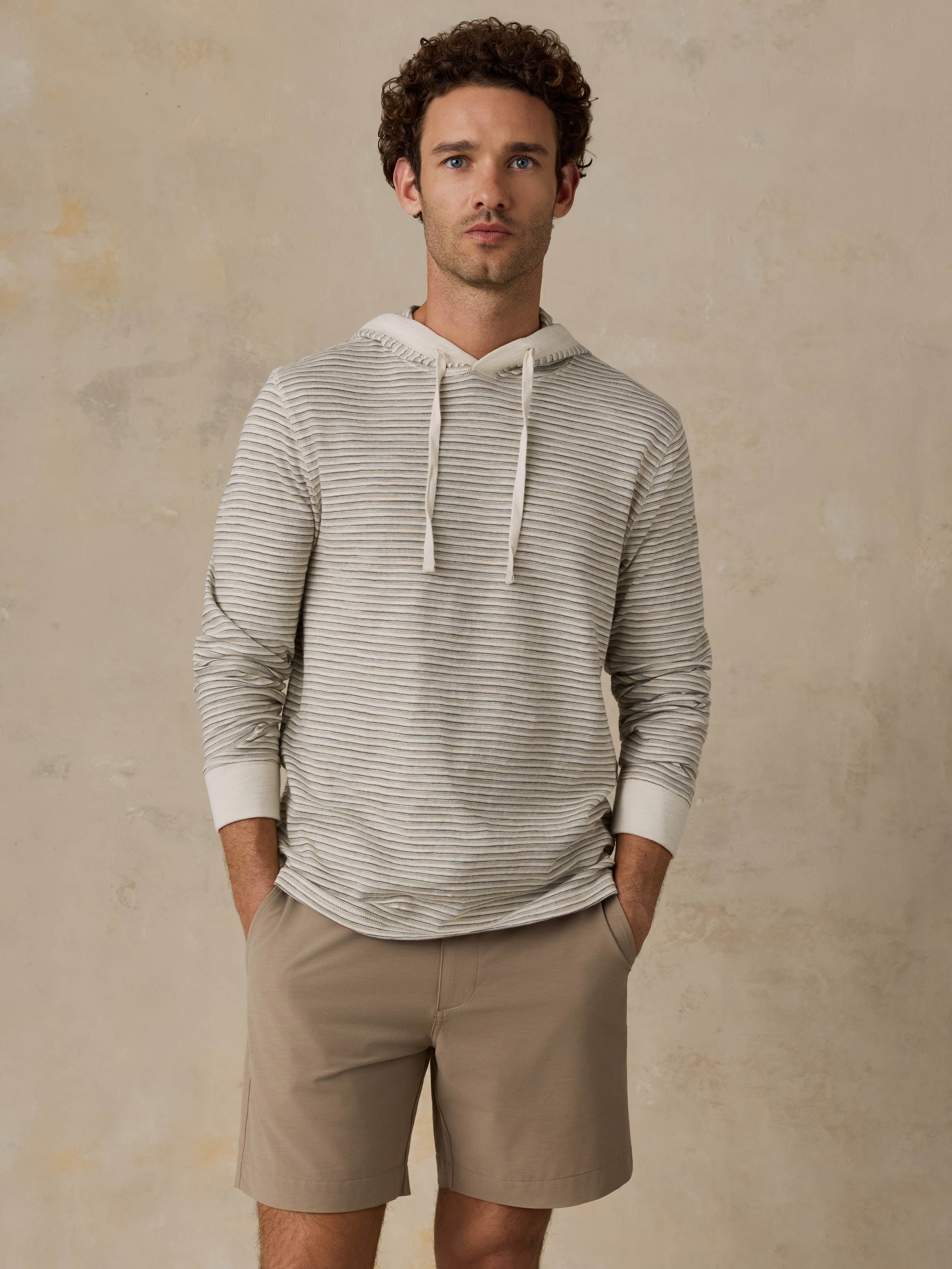 A man wearing a light beige and white horizontally striped long-sleeve hoodie with white drawstrings, paired with khaki shorts, standing against a textured beige studio backdrop.