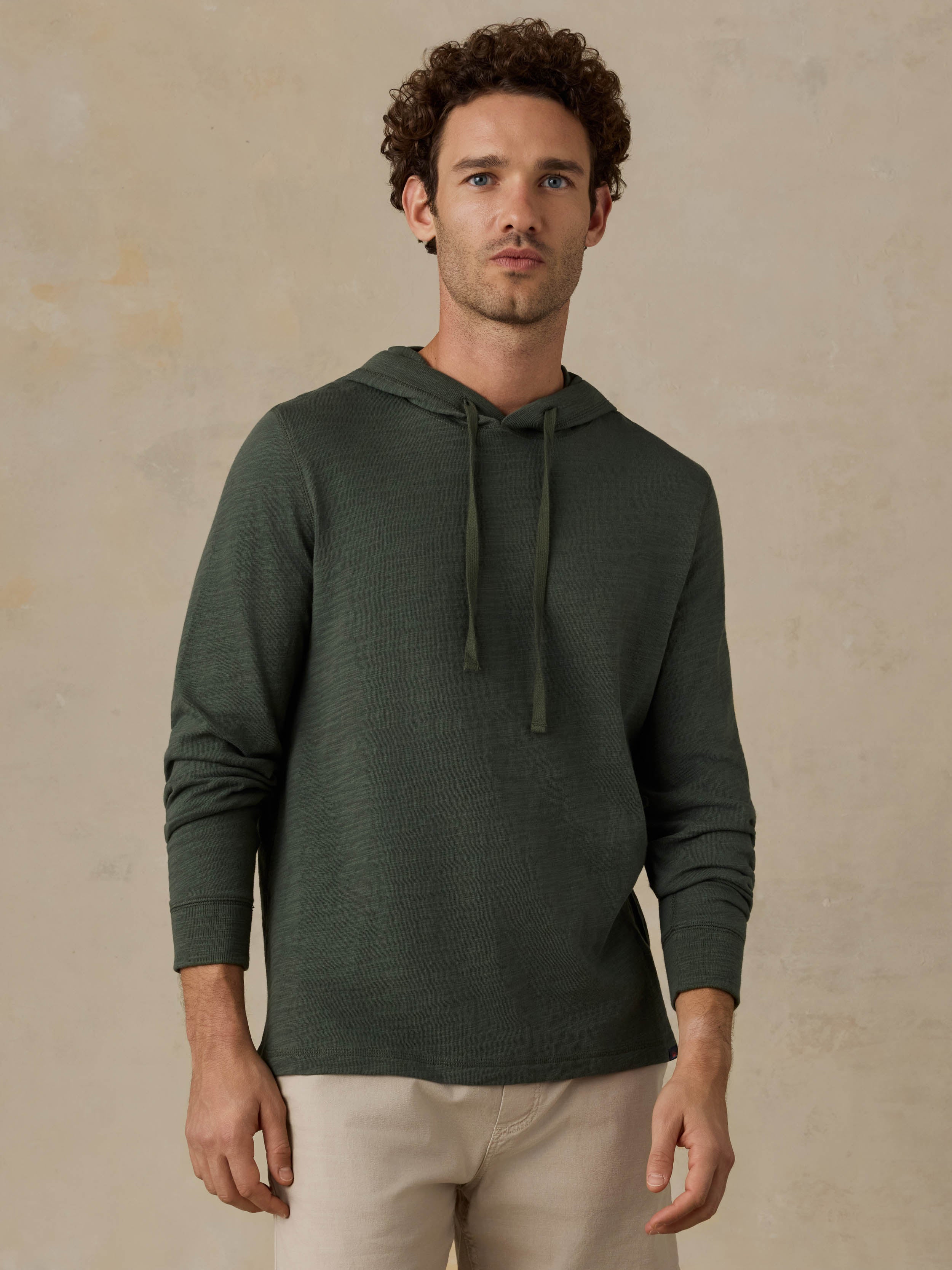 Man wearing a dark olive green hoodie and beige shorts against a beige background