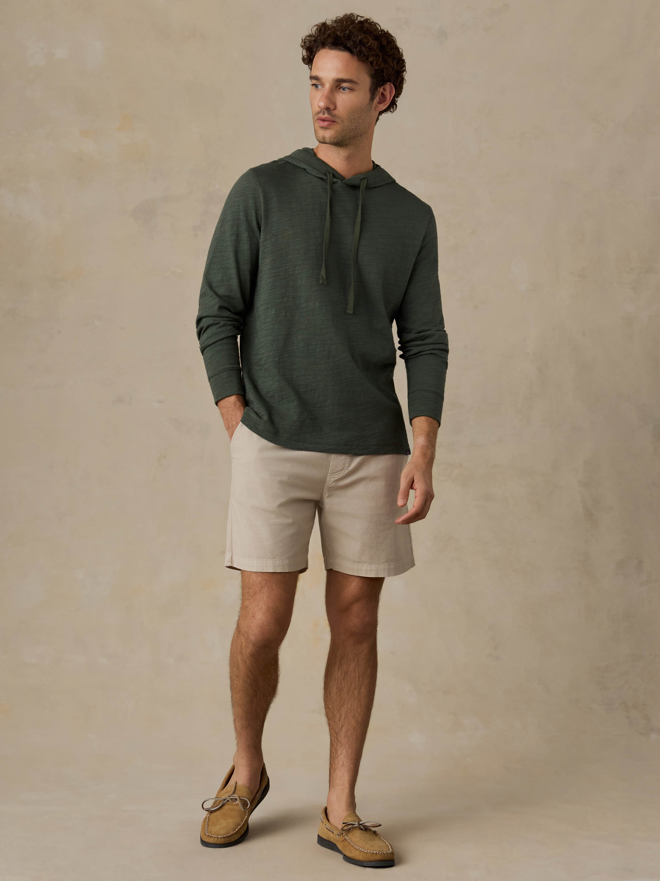 Man wearing a dark olive green hoodie, beige shorts, and boat shoes against a beige background