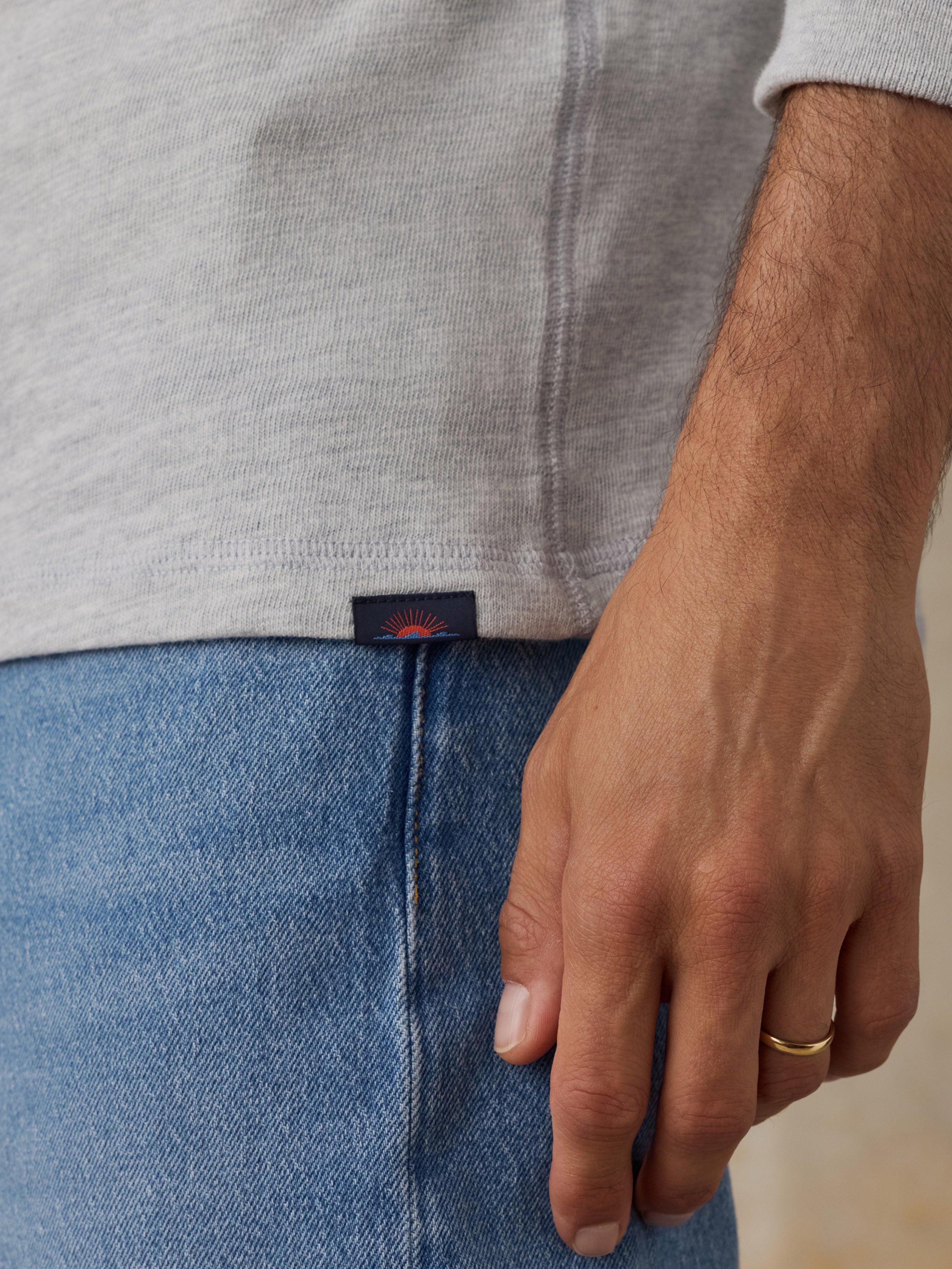 Close-up of a man wearing a light gray heathered knit top and blue jeans, with a small dark waistband label visible and a gold ring on the finger.