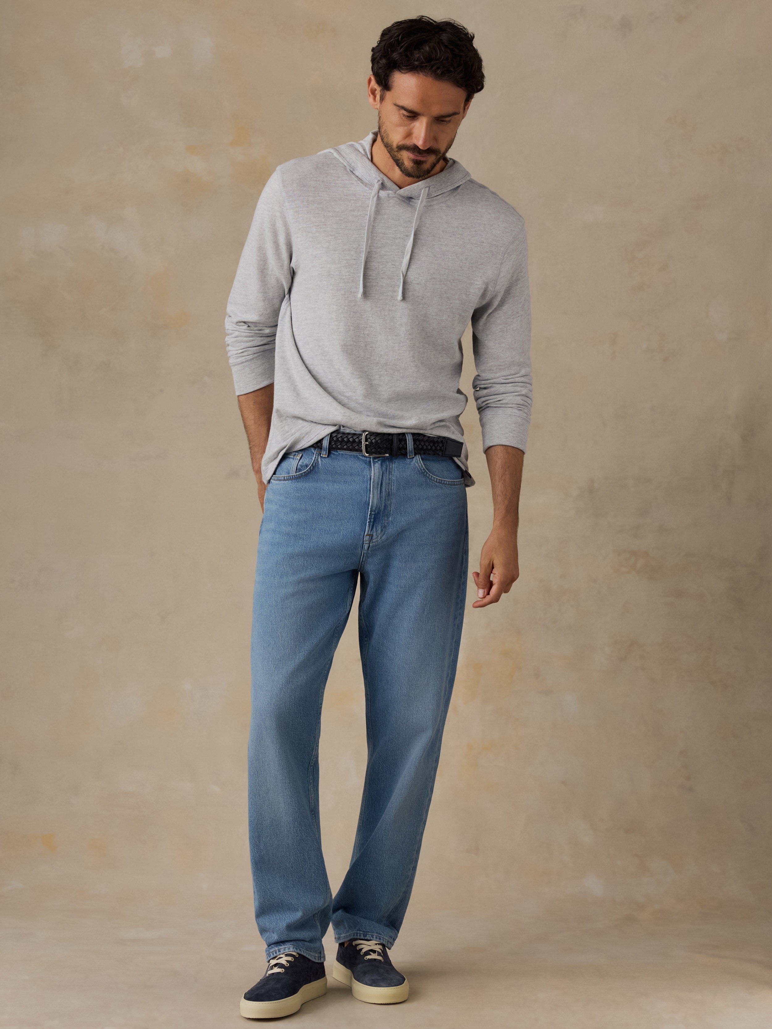 A man wearing a light gray long-sleeve hoodie, blue jeans, a black belt, and navy sneakers, standing against a textured beige studio backdrop.