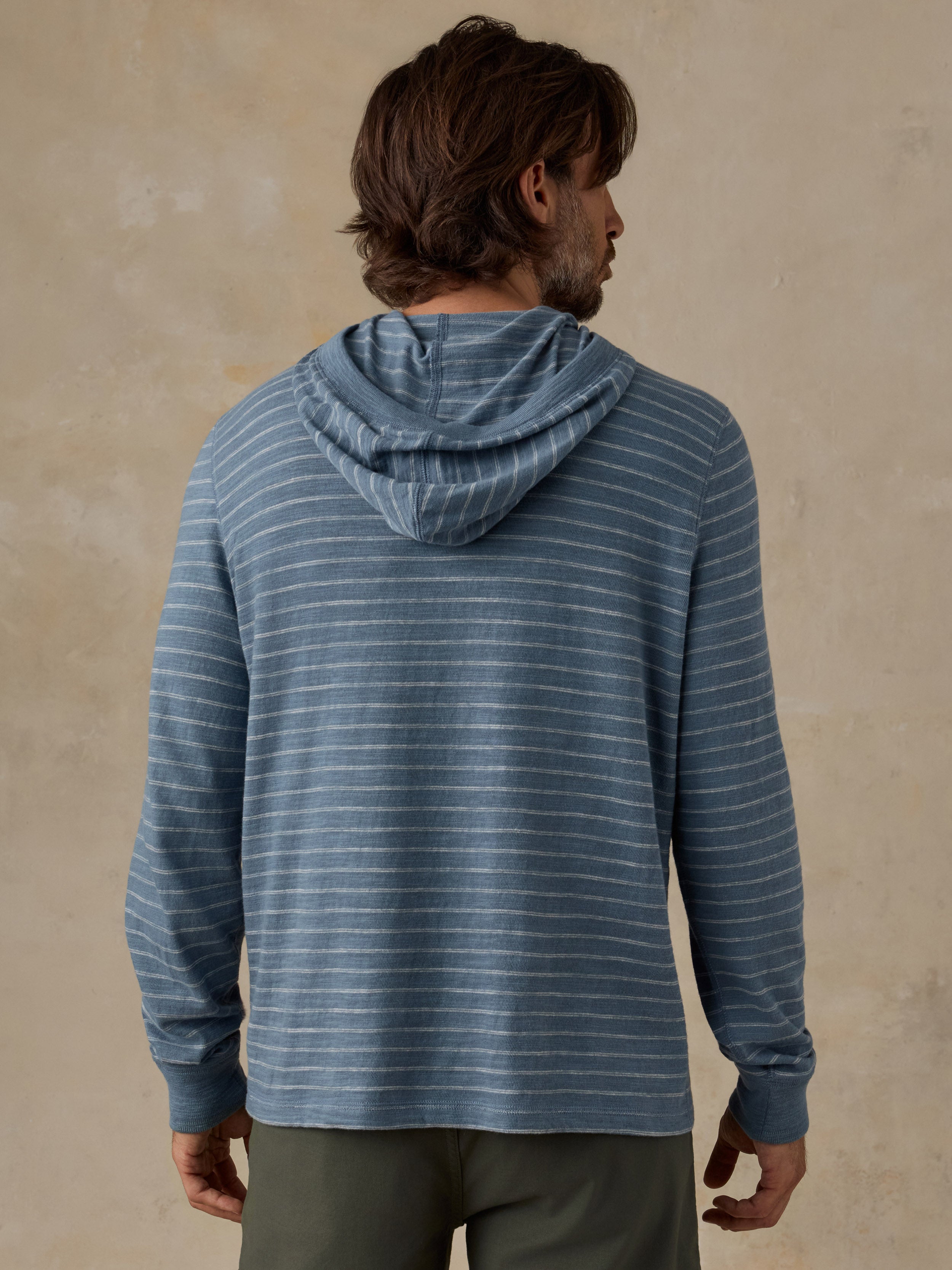 A man viewed from the back wearing a blue and white striped hoodie, set against a textured beige wall.