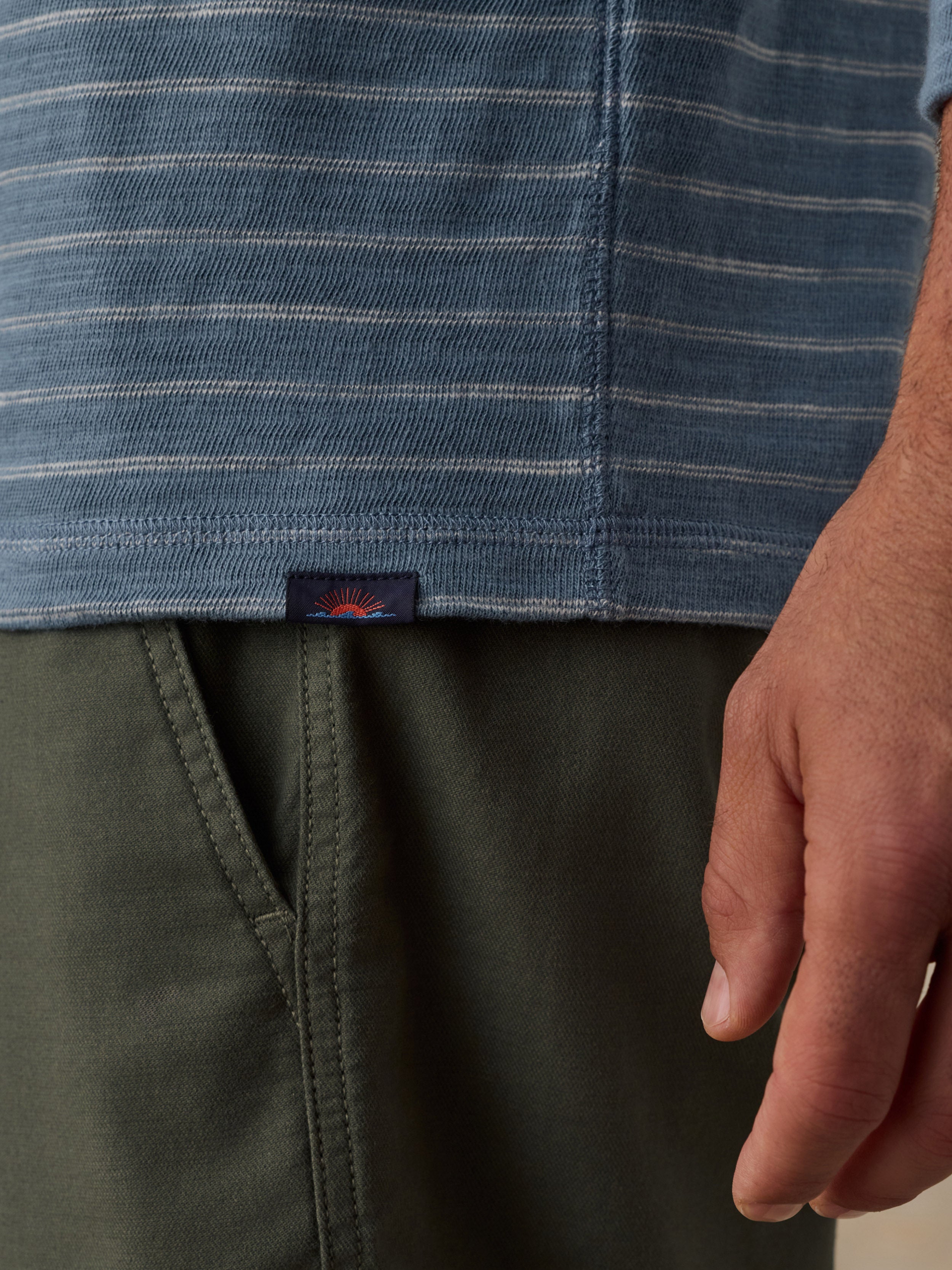Close-up of a man wearing a blue and white striped hoodie and olive green pants, with a small Faherty brand tab visible near the hem of the shirt.
