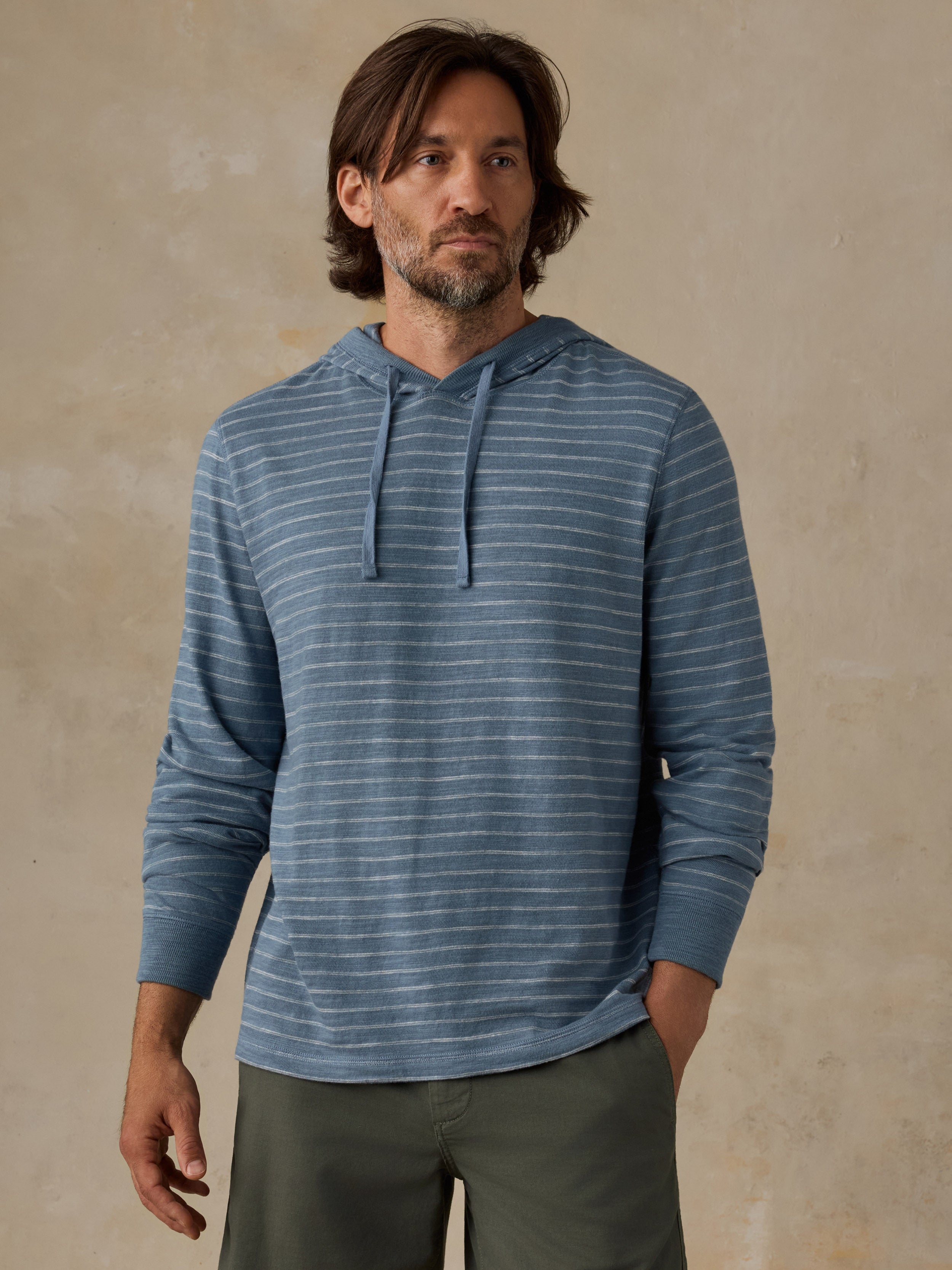 A man wearing a blue and white striped long-sleeve hoodie with olive green pants, standing against a textured beige wall.